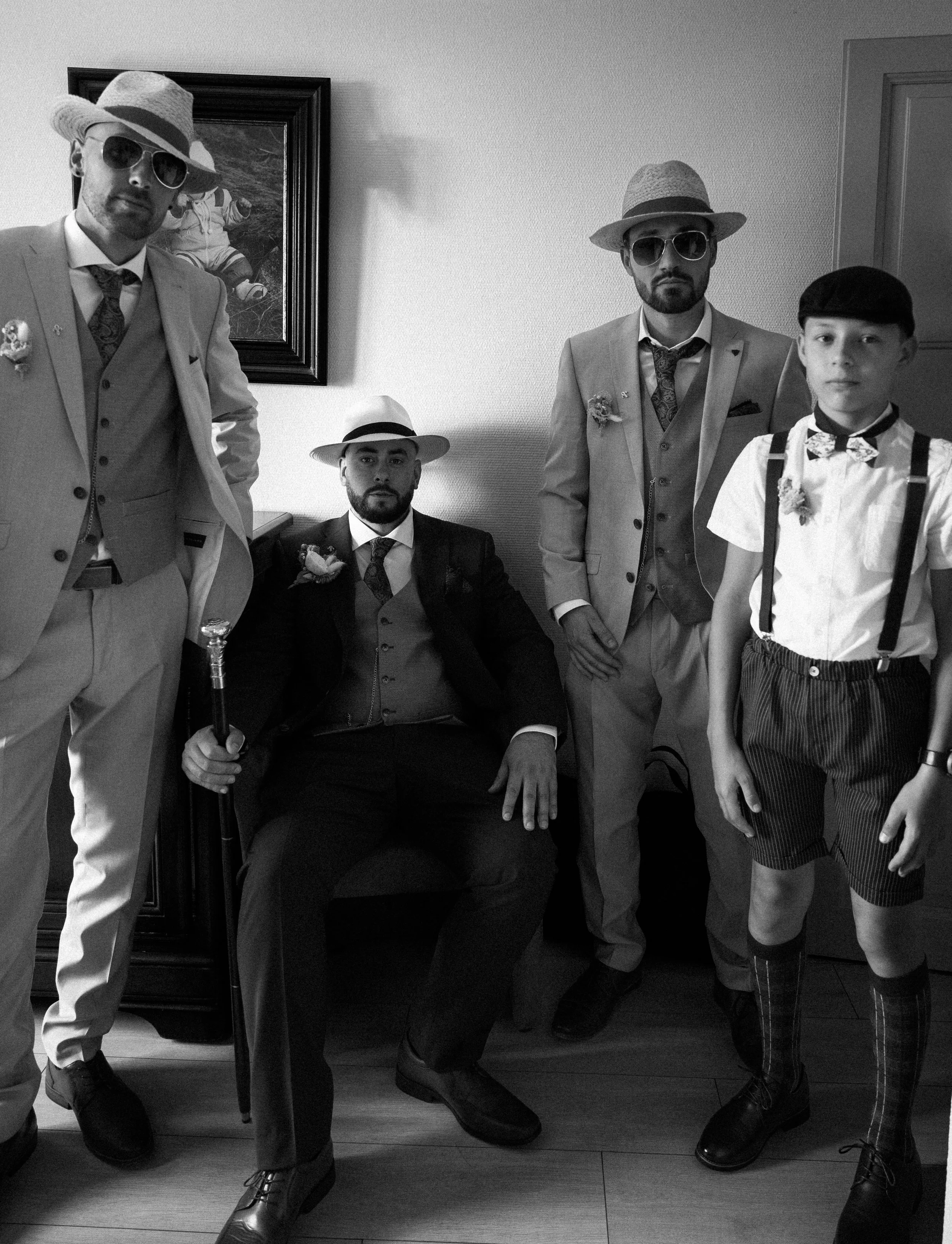 Cinq hommes posant dans une pièce, certains en costumes vintage et autres en vêtements modernes, avec accessoires comme chapeaux, lunettes, et sourire ou expression sérieuse.