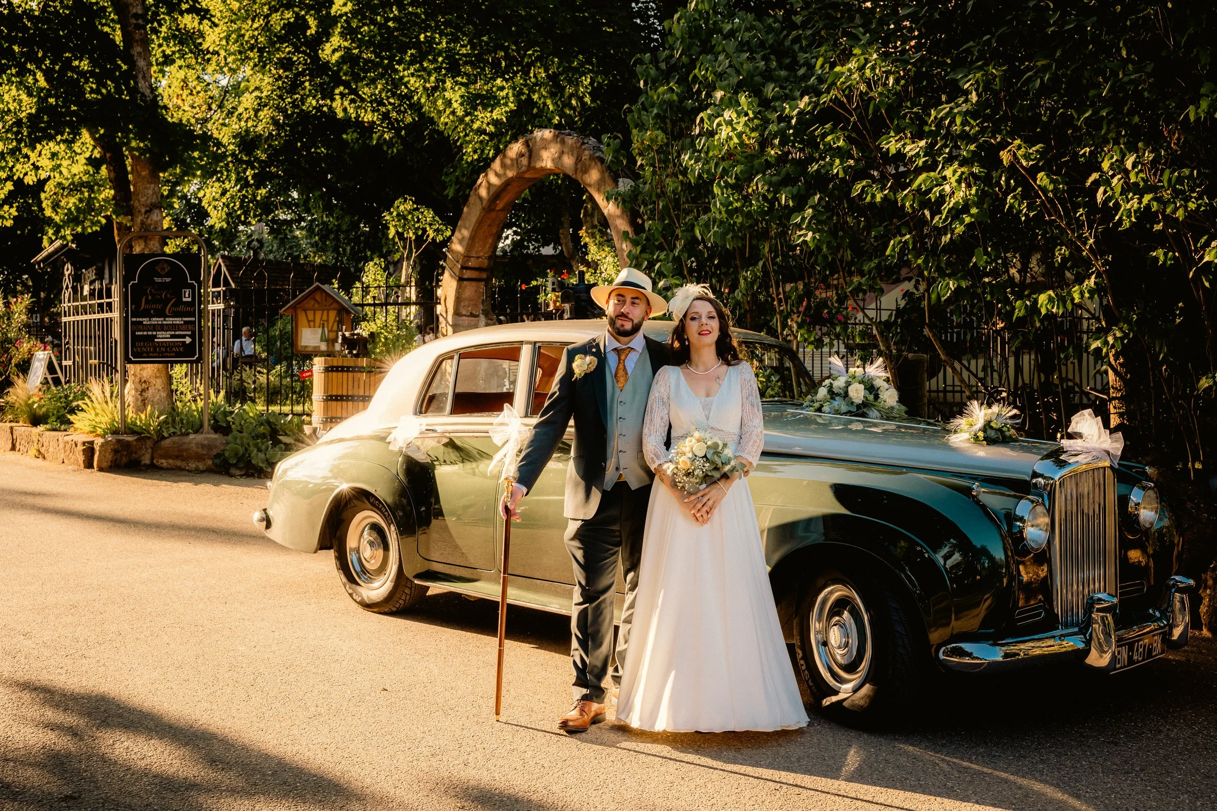 Un couple de mariés pose devant une voiture ancienne décorée pour un mariage, en plein jour, avec des arbres en arrière-plan.
