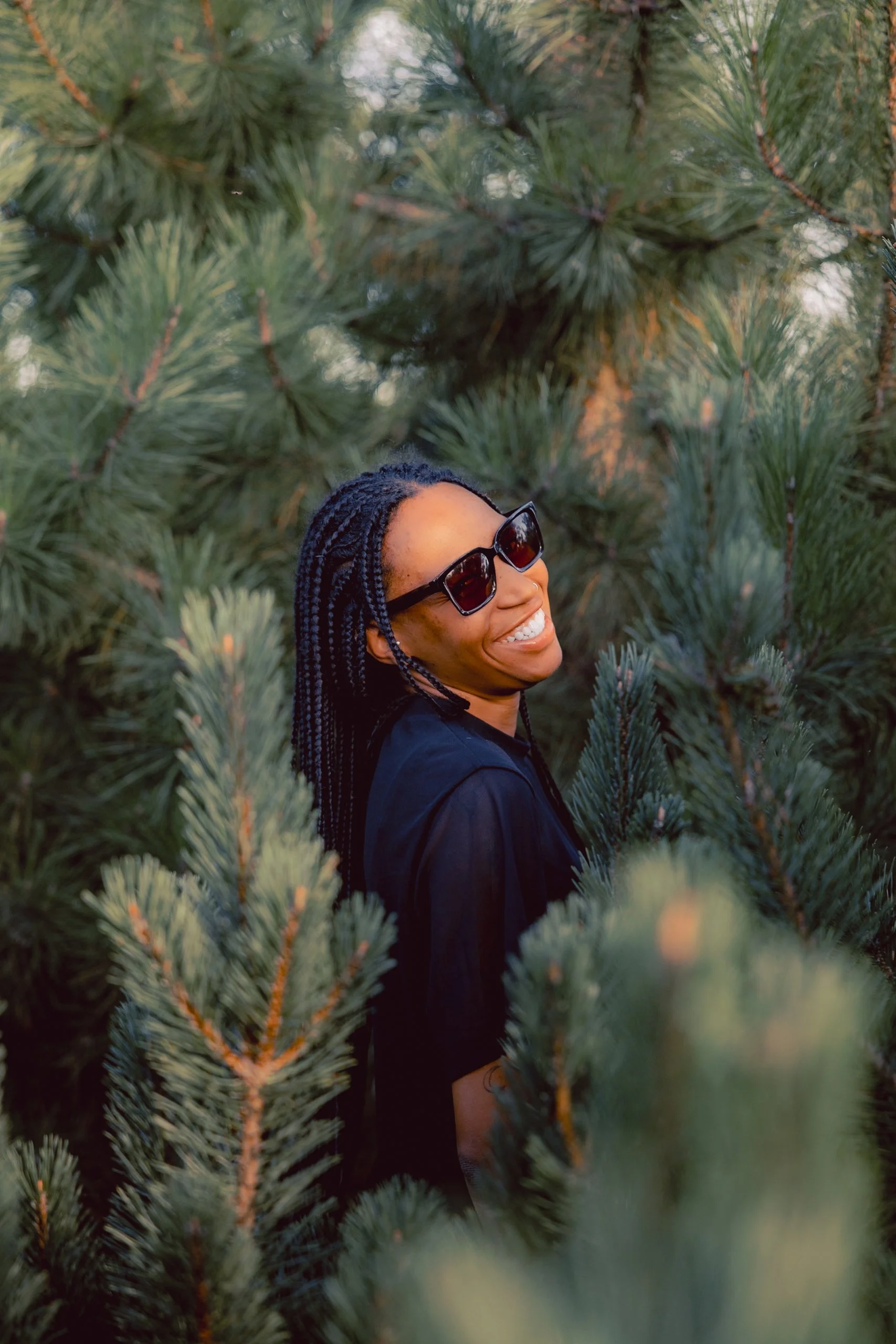 Femme souriante portant des lunettes de soleil, entourée d'arbres verts.