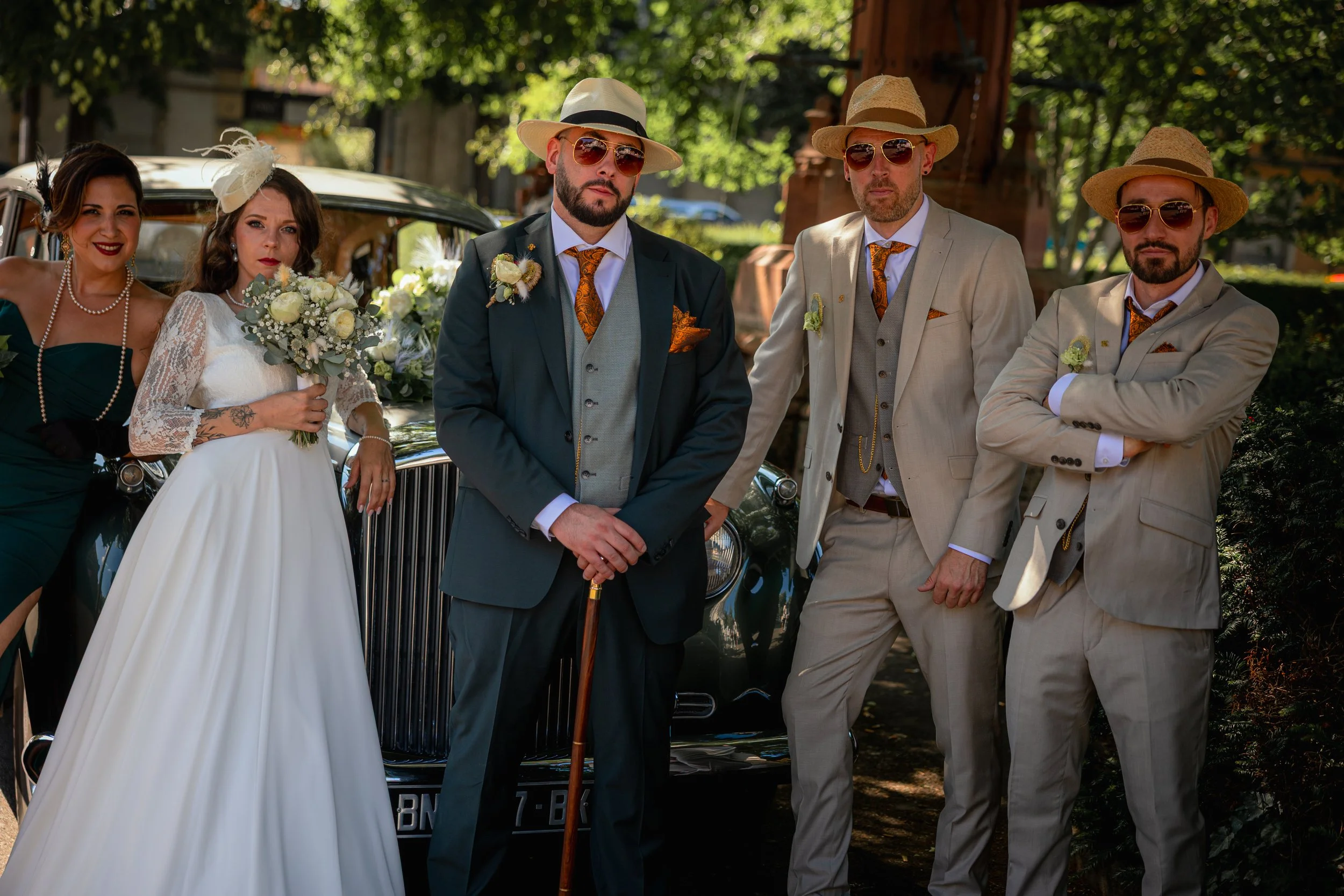 Groupe d'hommes et femmes lors d'un mariage, habillés en tenues élégantes, posant devant une voiture ancienne décorée, avec un peu de végétation en arrière-plan.