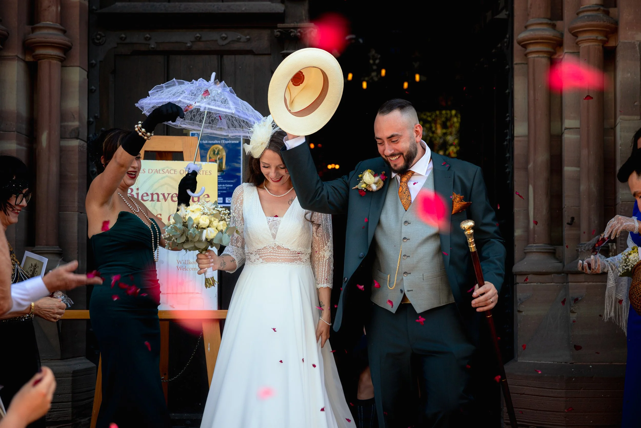 Un couple de mariés souriants, la mariée en robe blanche et le marié en costume, sortant de l'église sous une pluie de pétales de rose, entourés de leurs invités.