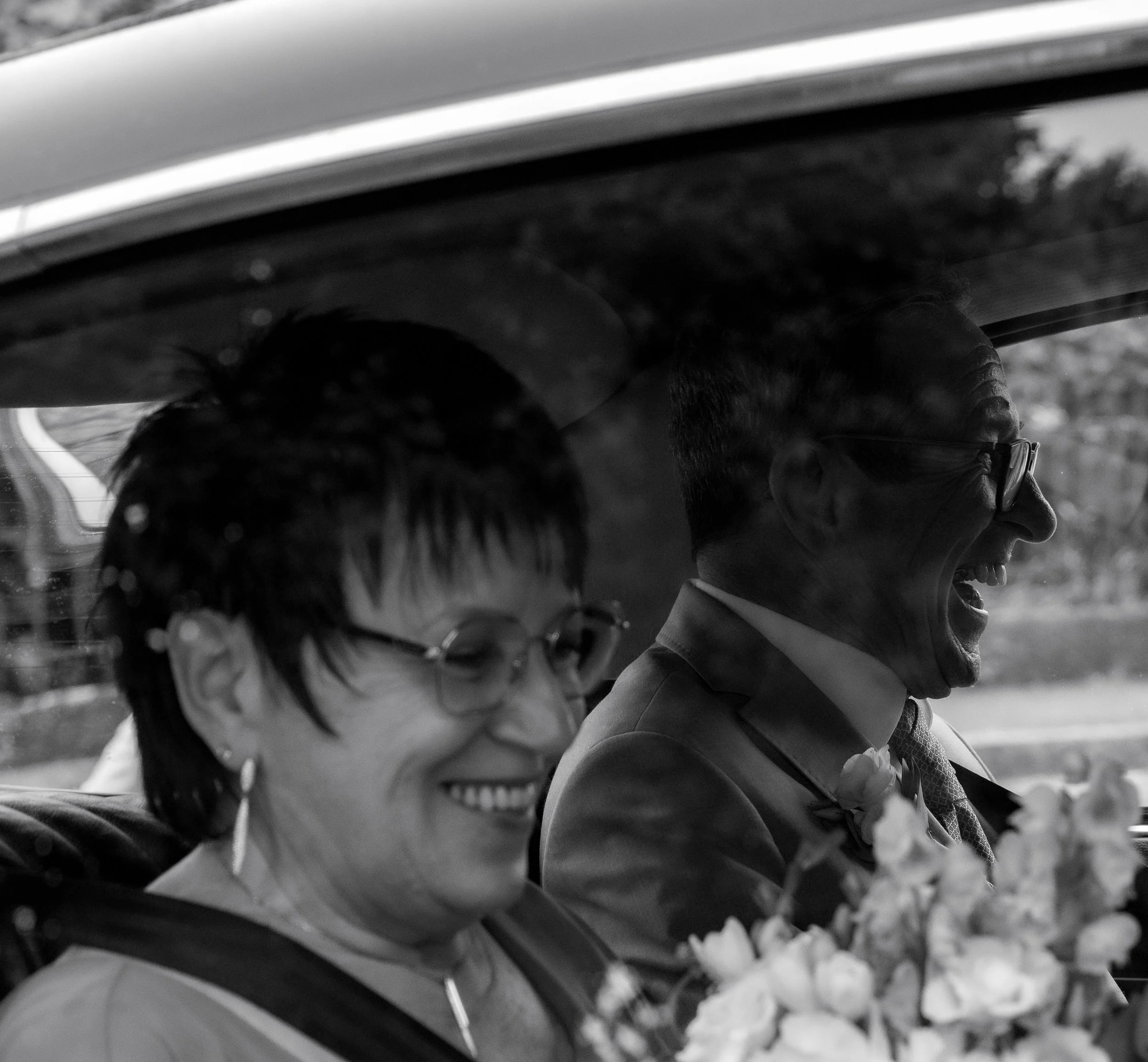 Un couple souriant assis dans une voiture, probablement lors d'une occasion spéciale, comme un mariage, avec des fleurs devant eux.