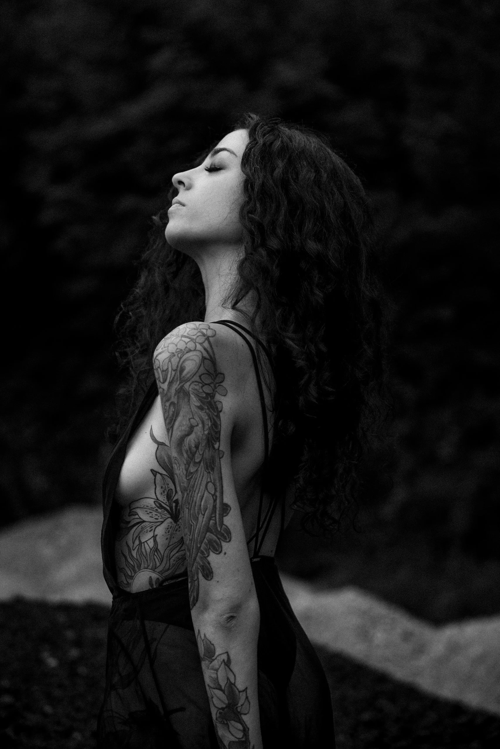 Une femme avec des tatouages sur le bras, les épaules et l'épaule, les yeux fermés, en profil, dans un environnement sombre.