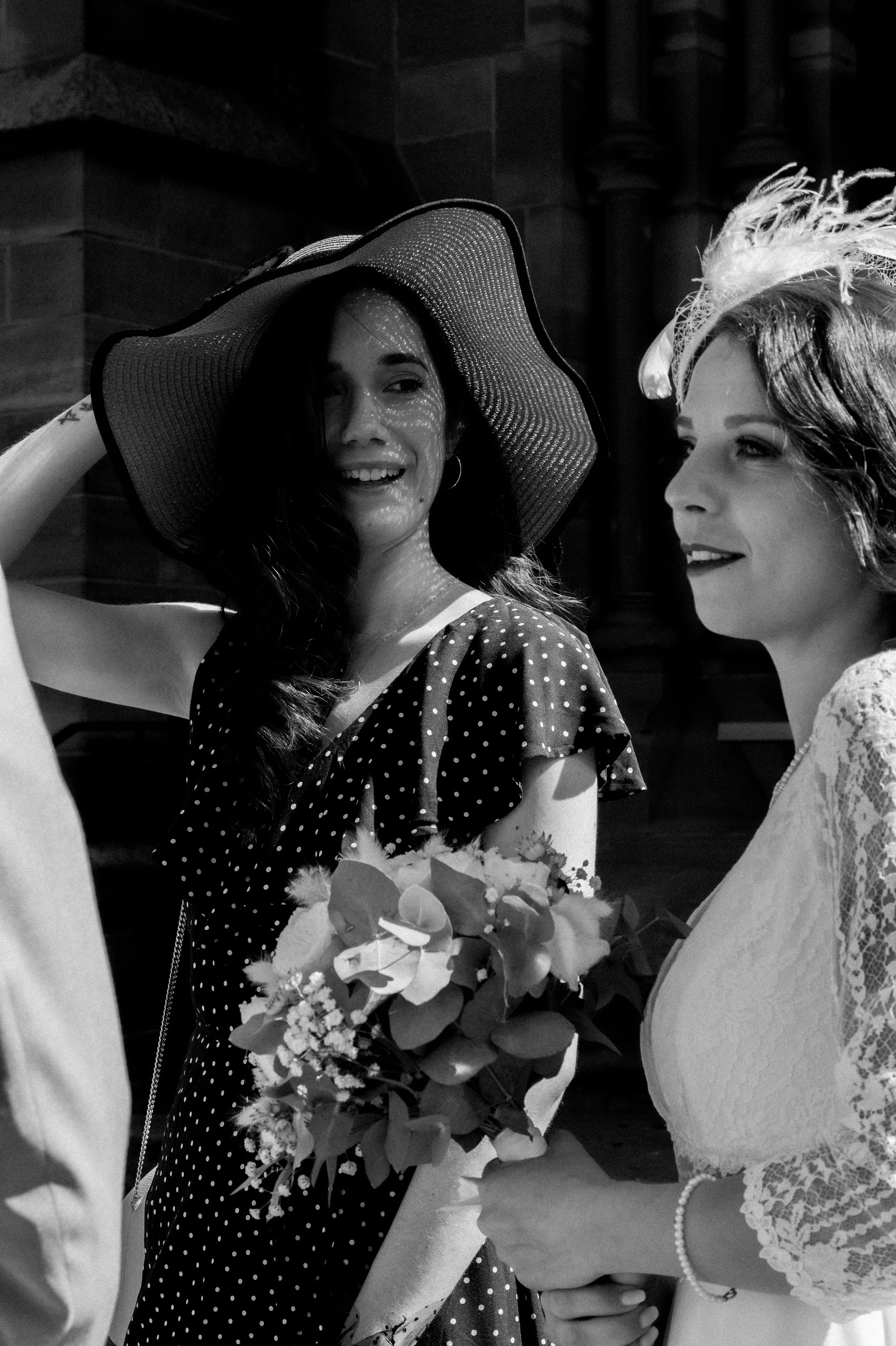 Deux femmes lors d'une célébration, l'une porte un grand chapeau à large bord et tient un bouquet de fleurs, l'autre porte une tenue en dentelle avec un peigne dans les cheveux.