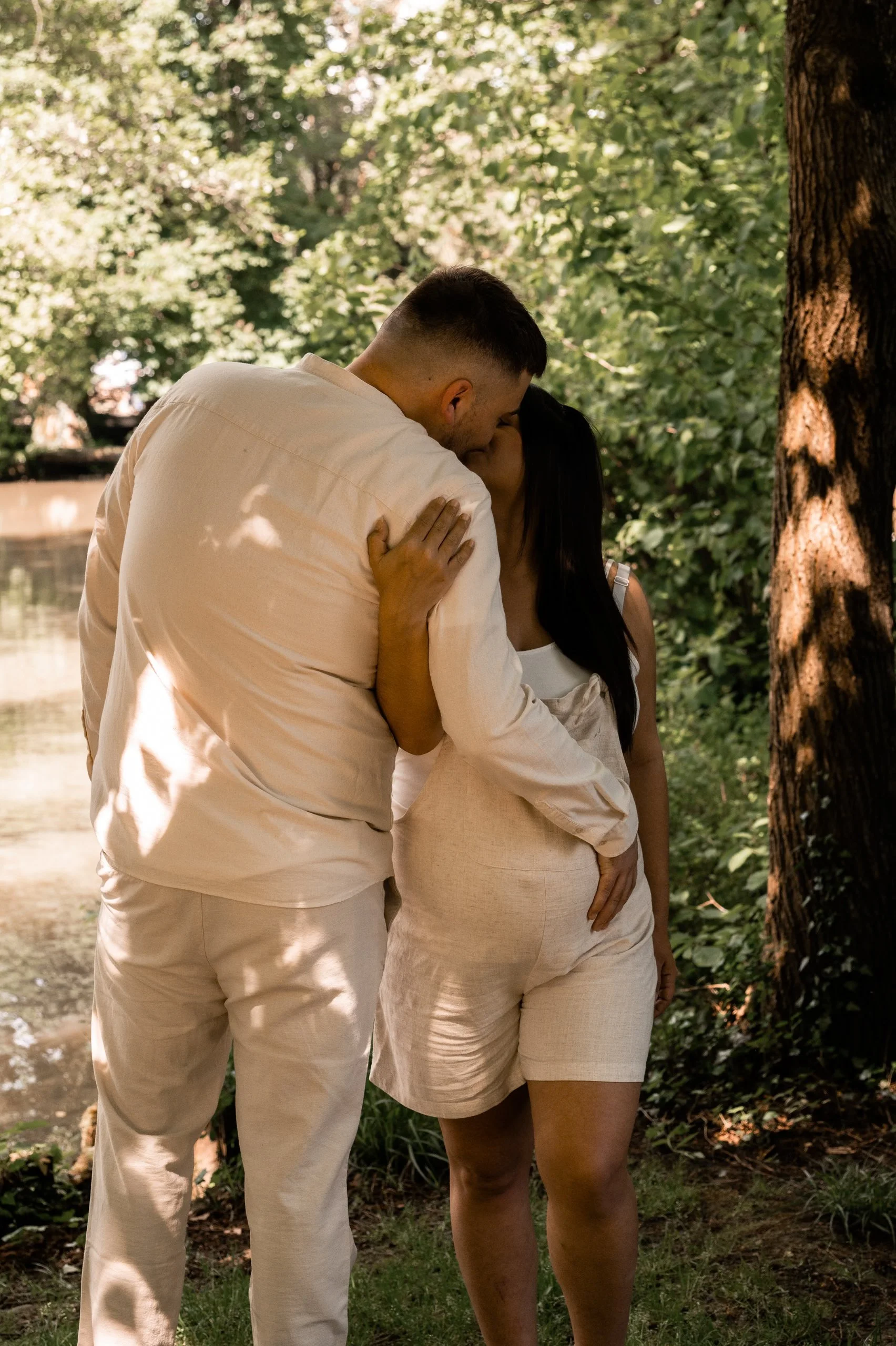 Un couple s'embrassant dans un parc, entouré d'arbres verts et d'une rivière en arrière-plan.