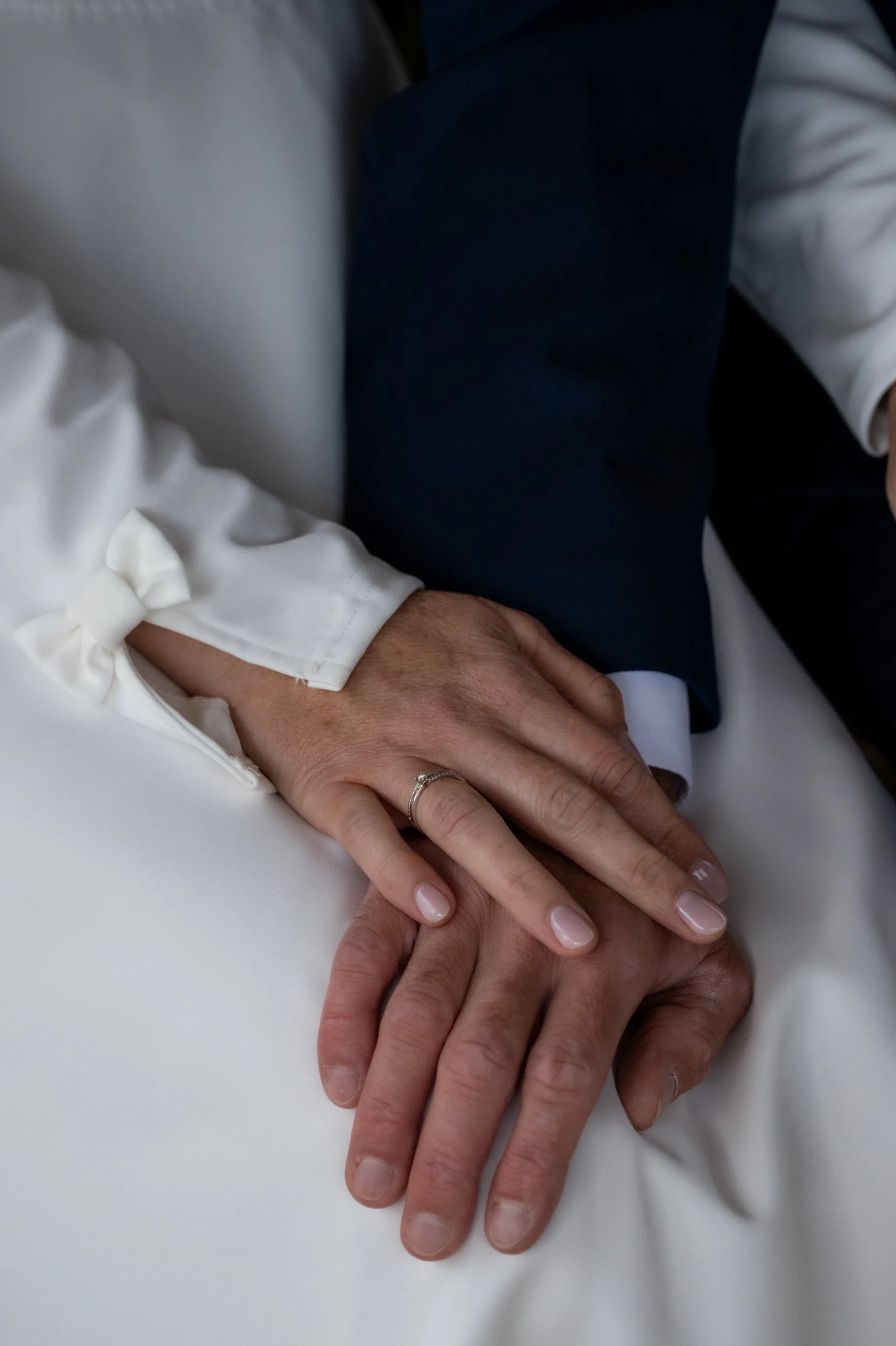 Mains croisées de deux personnes, une porteuse d'une alliance, posées sur un drap blanc.