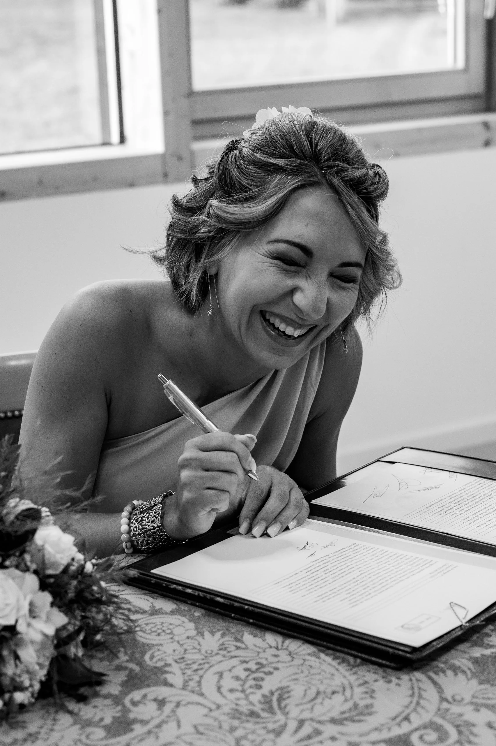Une femme qui sourit en signant un document, probablement lors d'une cérémonie, en noir et blanc.