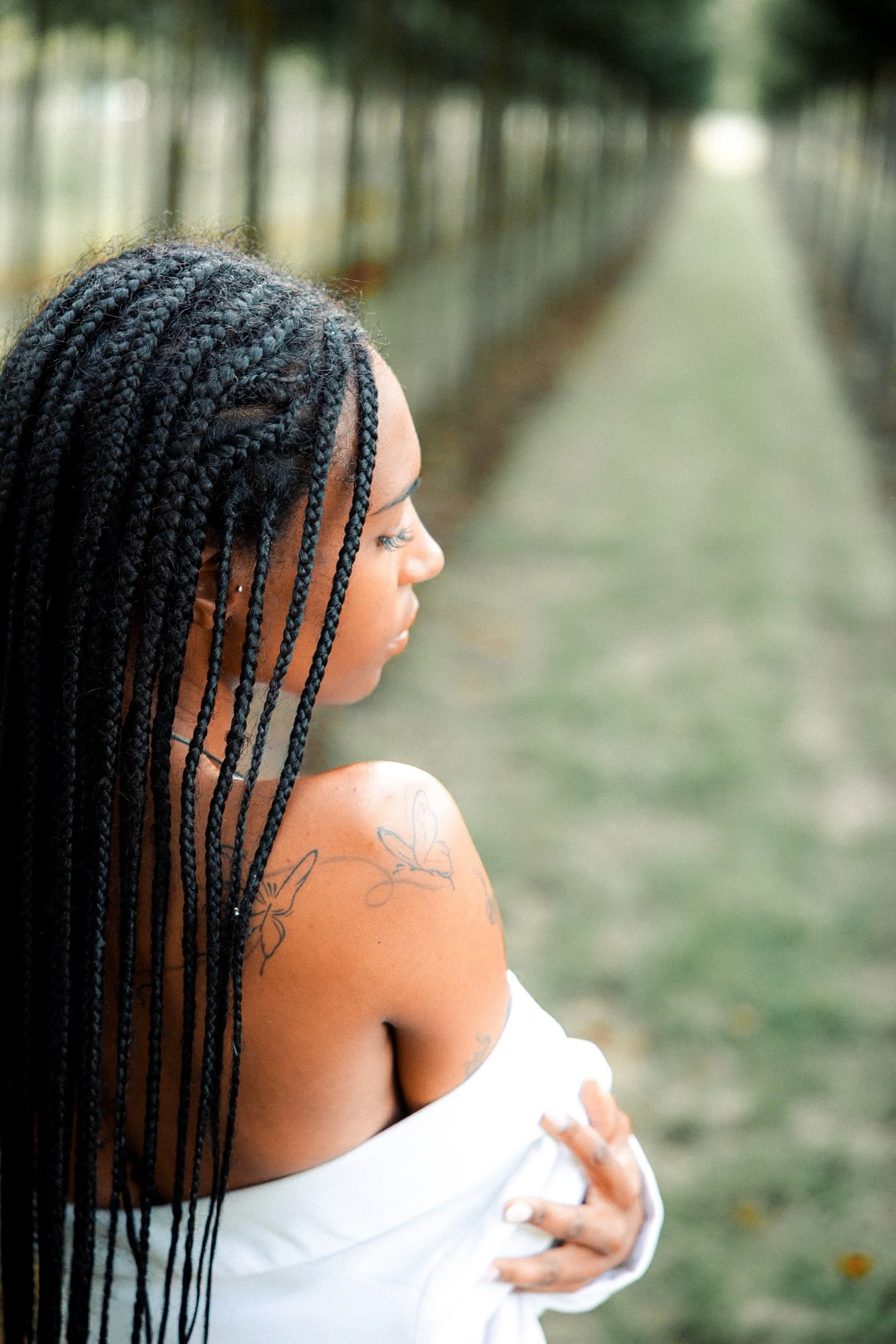 Femme avec des cheveux en tresses portant un haut blanc, debout dans un lieu extérieur avec un chemin bordé d'arbres.
