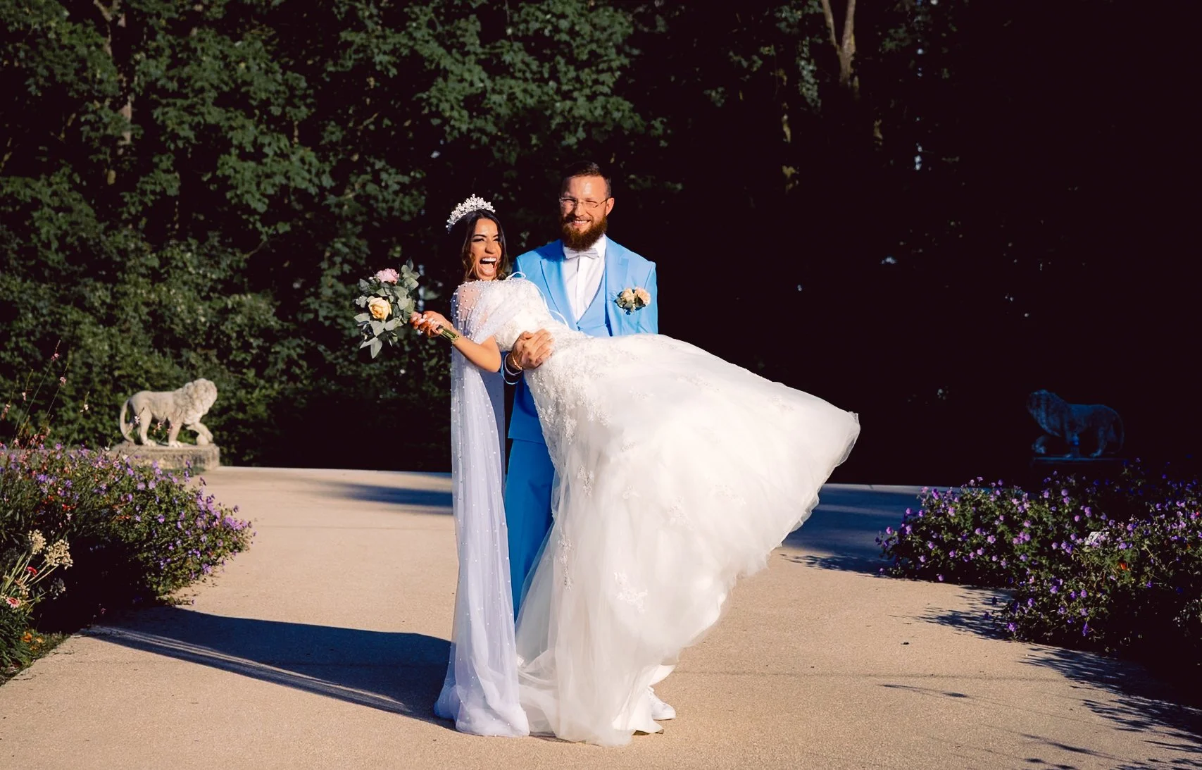 Un couple de mariés souriants, la femme en robe de mariée blanche portant un bouquet et le marié en costume bleu clair, posant en extérieur à l'ombre, avec deux sculptures d'animaux en pierre dans un jardin fleuri.