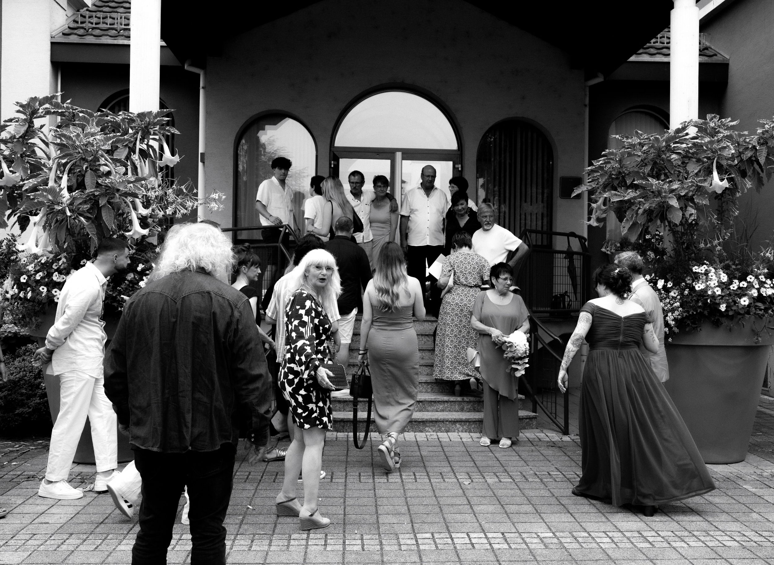 Groupe de personnes rassemblées devant un bâtiment, probablement pour un événement ou une cérémonie, avec des fleurs en décoration.