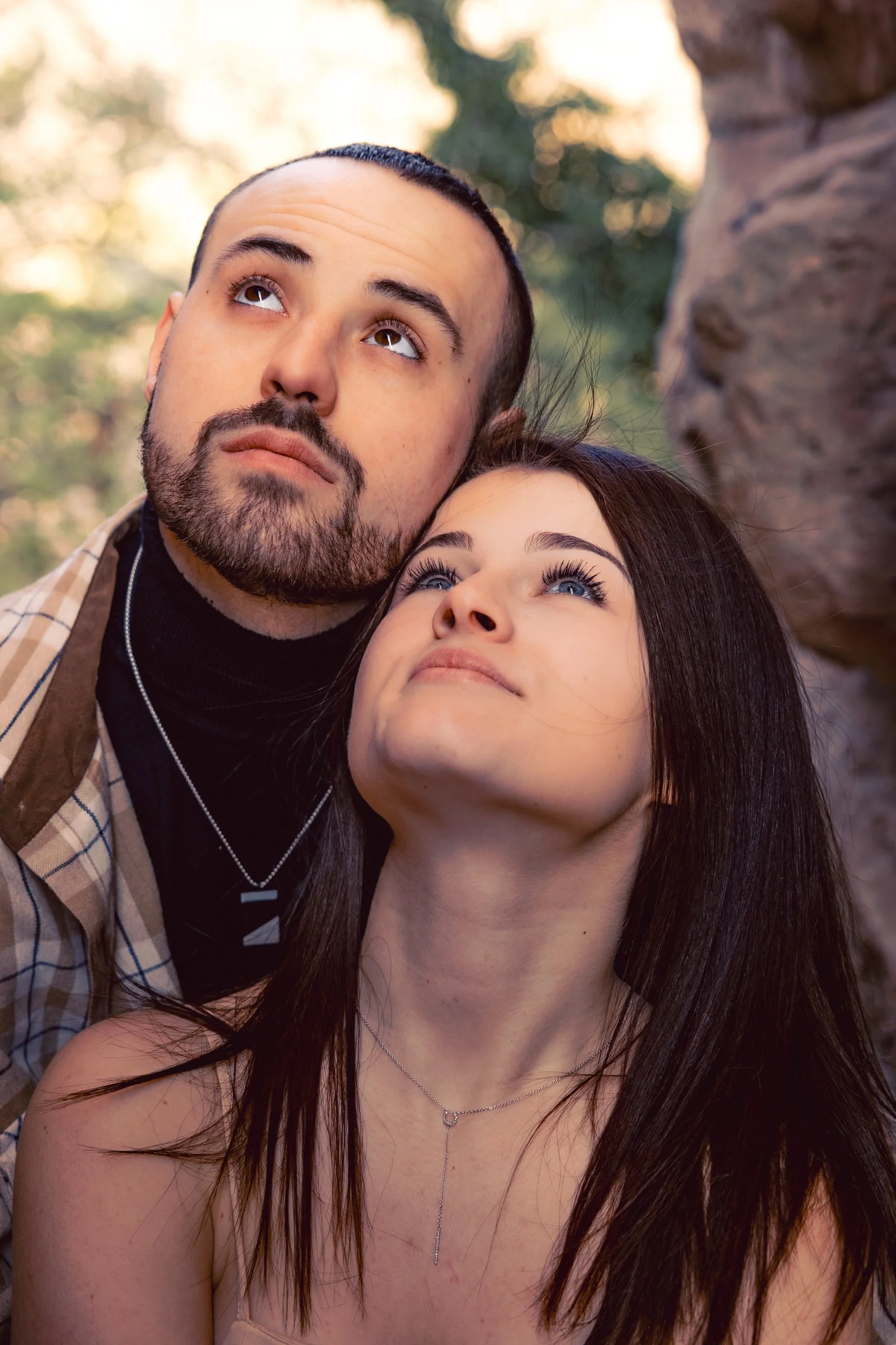 Un jeune homme et une jeune femme regardant vers le haut avec une expression contemplative, en extérieur avec des arbres en arrière-plan.