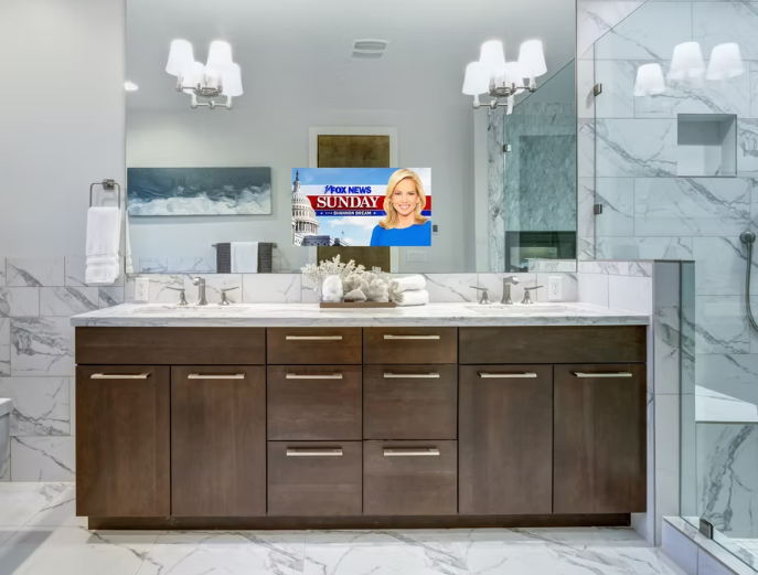 Modern bathroom with a large mirror, wooden vanity with drawers, marble countertop, and wall-mounted faucet. A TV is mounted on the wall facing the sink, displaying a Fox News program with a woman and Capitol building in the background.