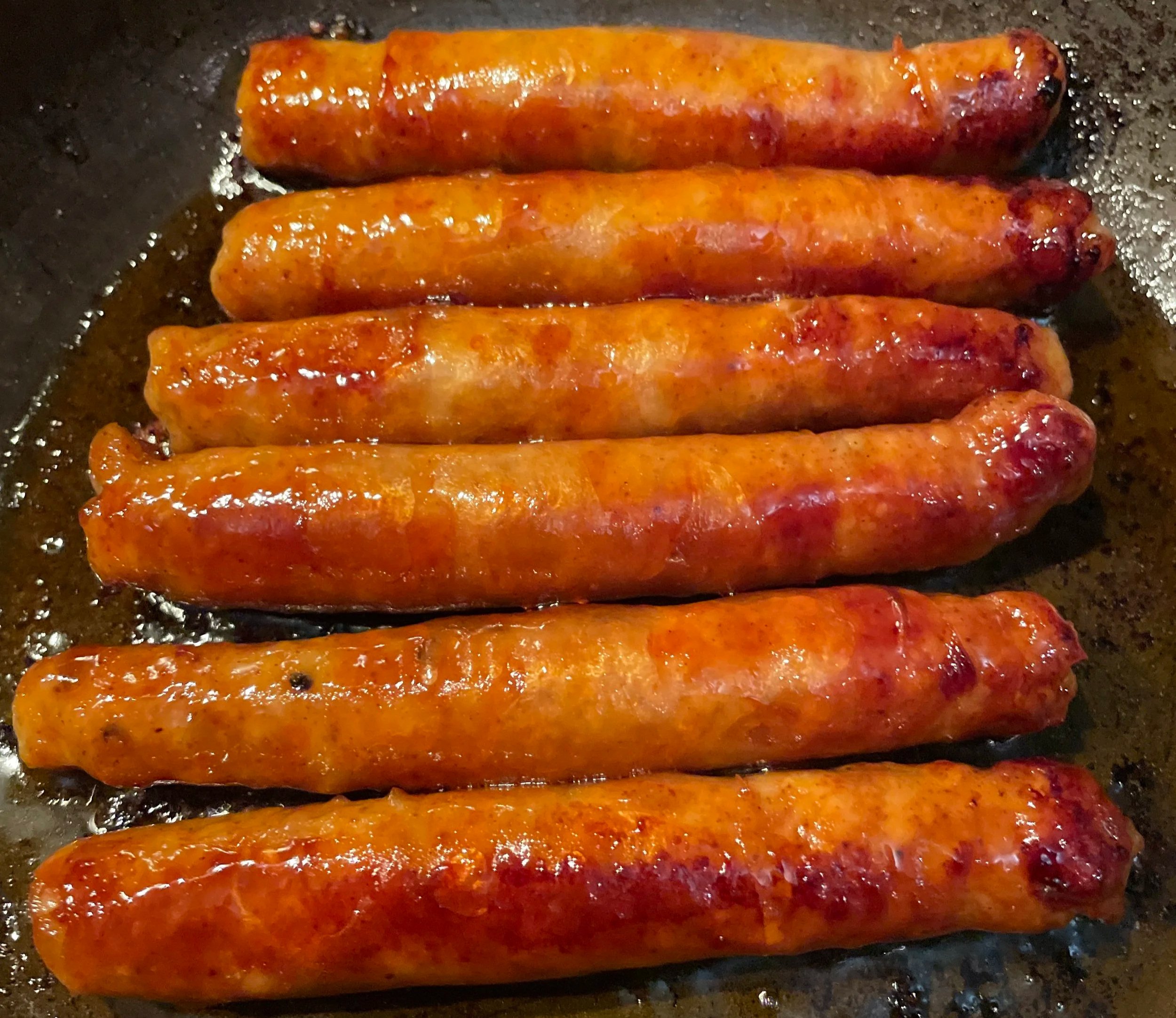 Seven browned sausages frying in a black skillet.