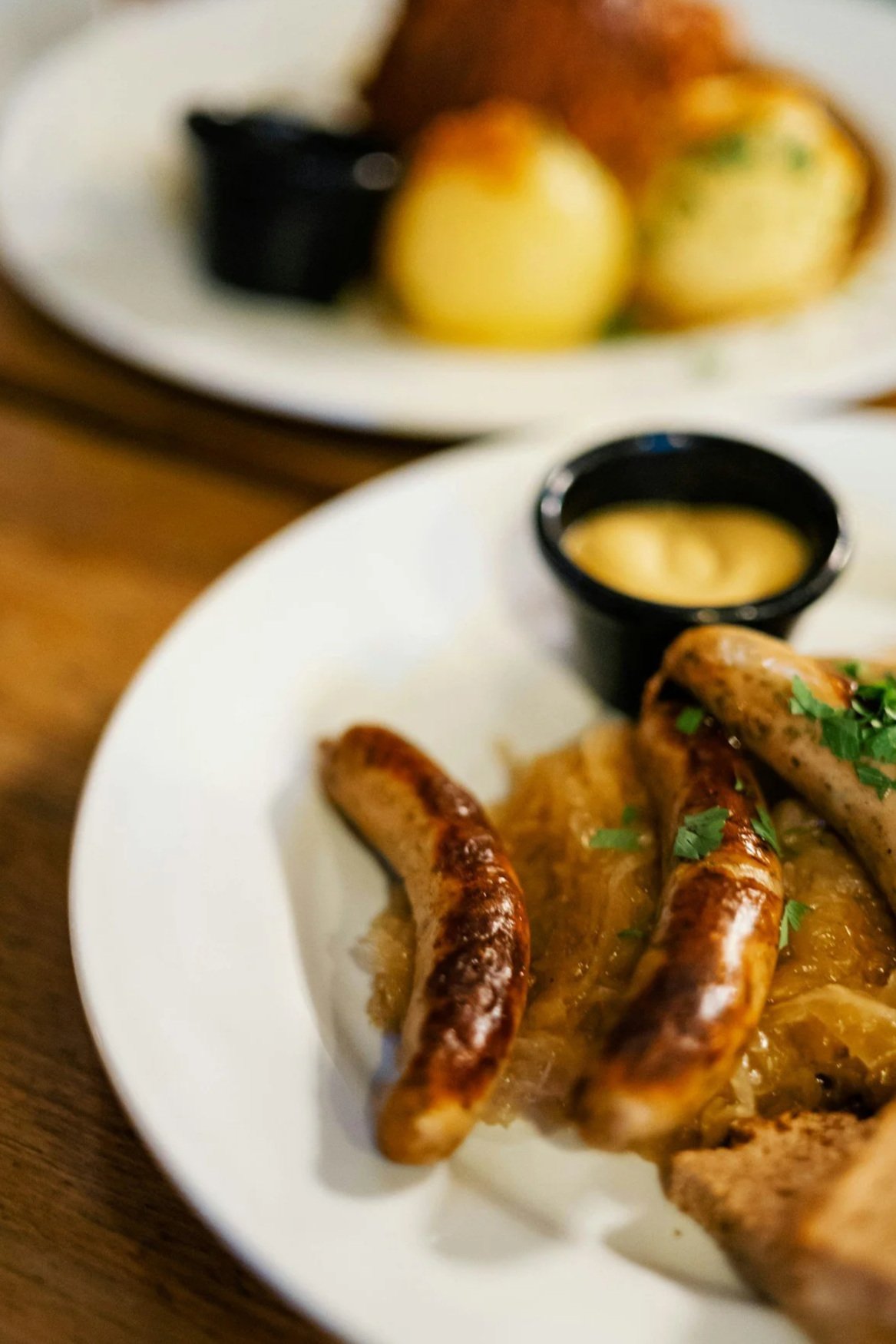 Close-up of sausages with caramelized onions and fresh herbs on a white plate, served with a side of mustard in a small black cup, and a separate dish with potatoes and possibly meat in the background.