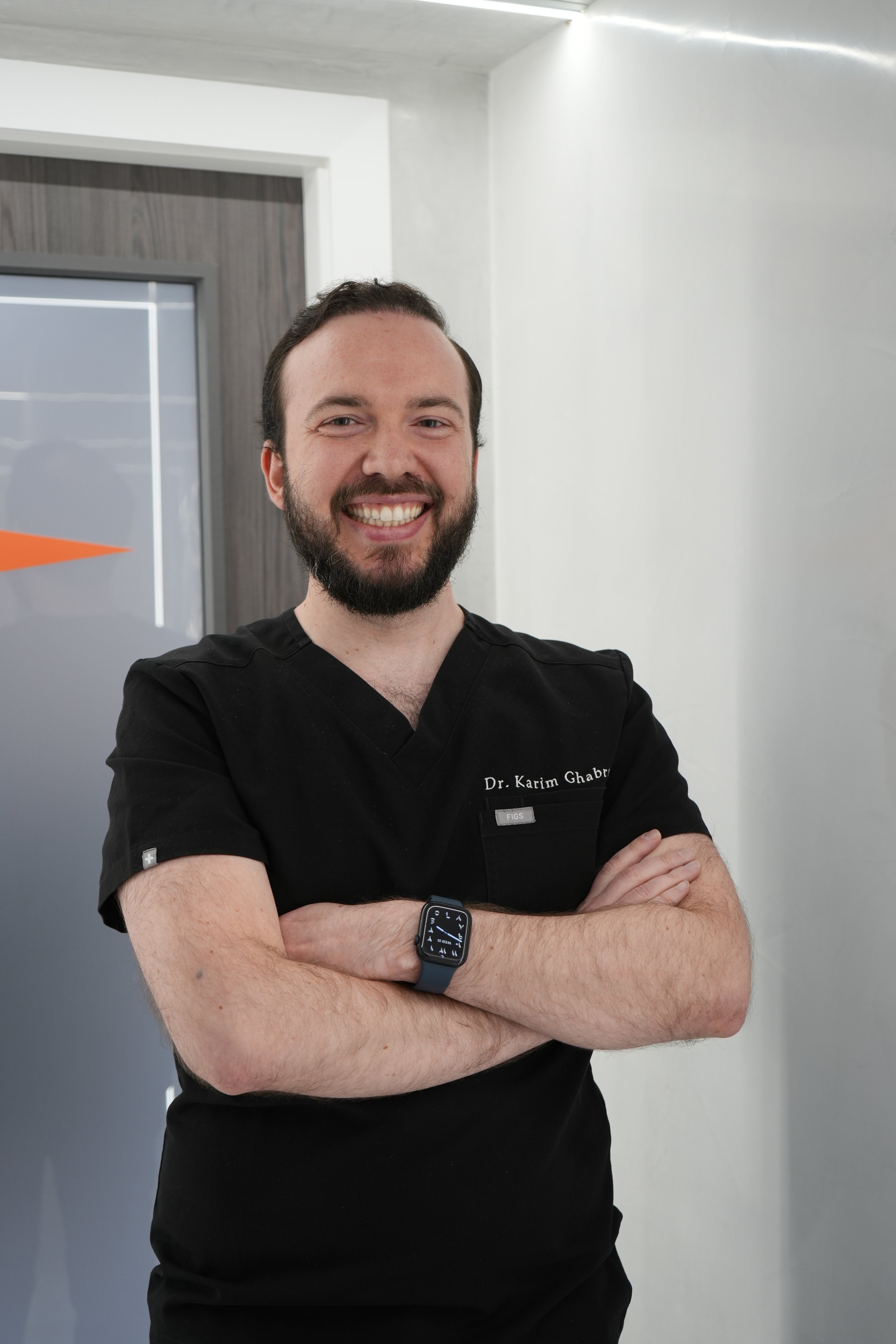 A smiling man with a beard and short dark hair, wearing black medical scrubs and a smartwatch, standing with arms crossed in a clinic or hospital setting.
