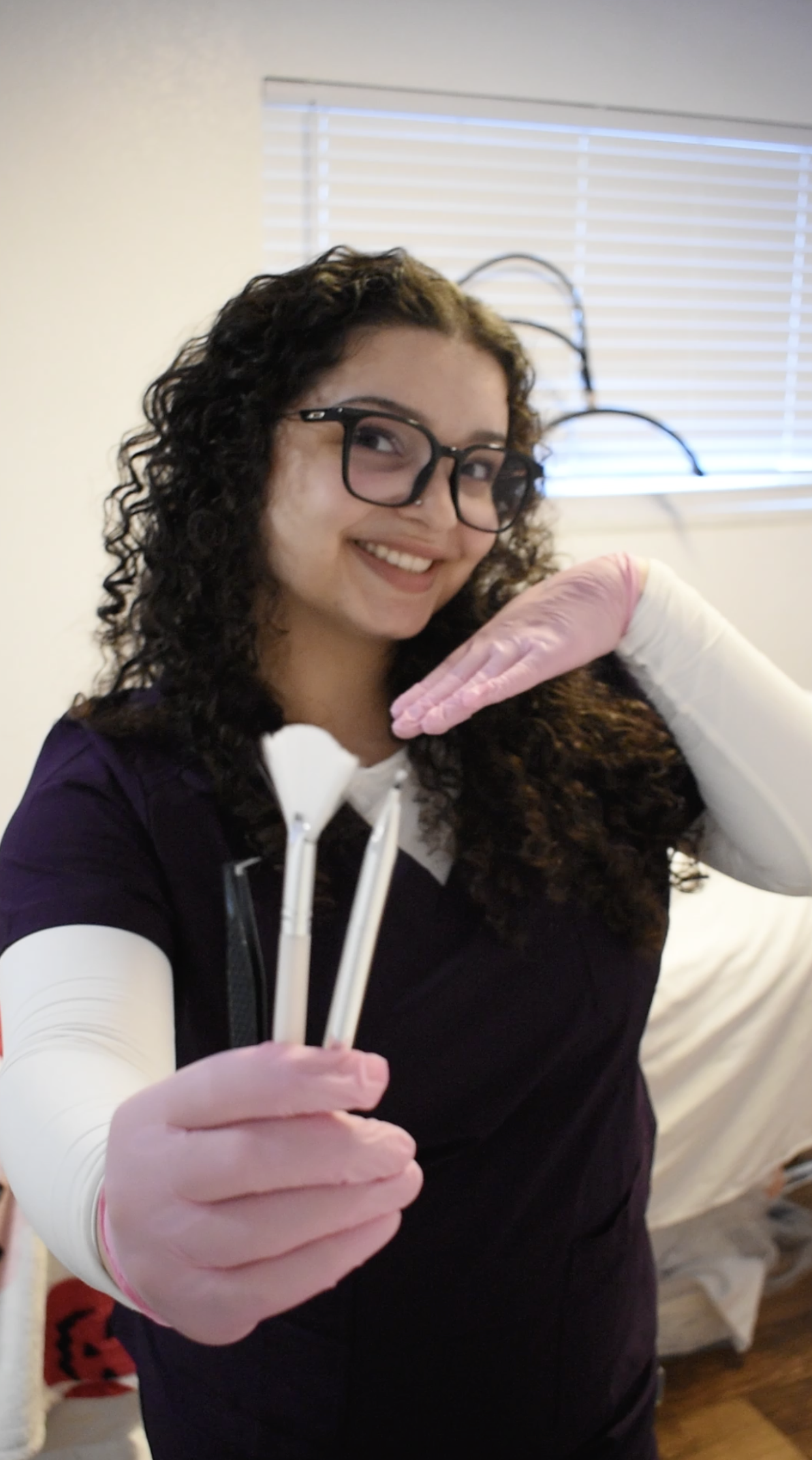 Smiling woman in glasses and pink gloves holding implements like facial mask brush and eyebrow tinting brush