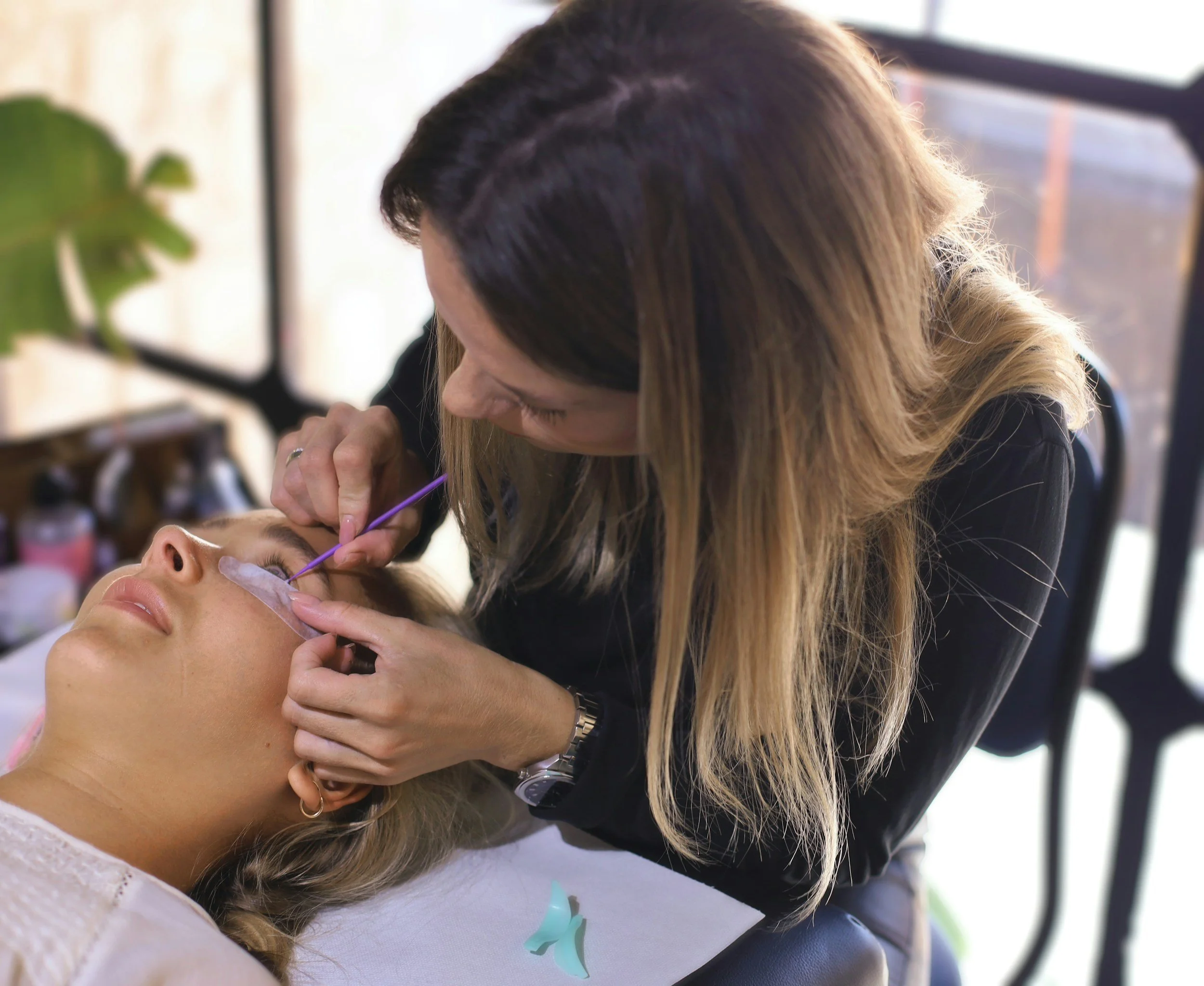 A woman with blonde hair receiving eyelash extensions from a professional technician in a salon.