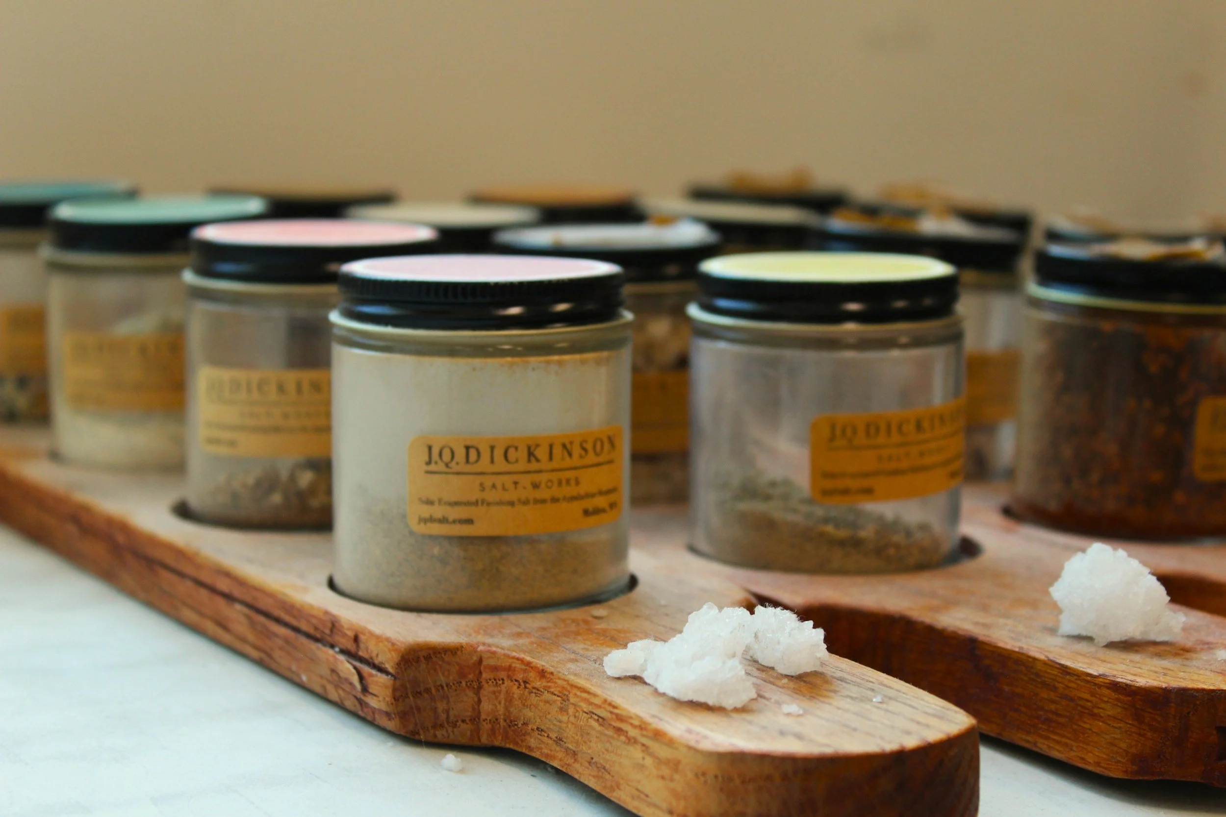 Multiple jars of salt with black lids and labels that read "J.Q. Dickinson Salt Works" are arranged on a wooden board. Small piles of salt are scattered in front of the jars.