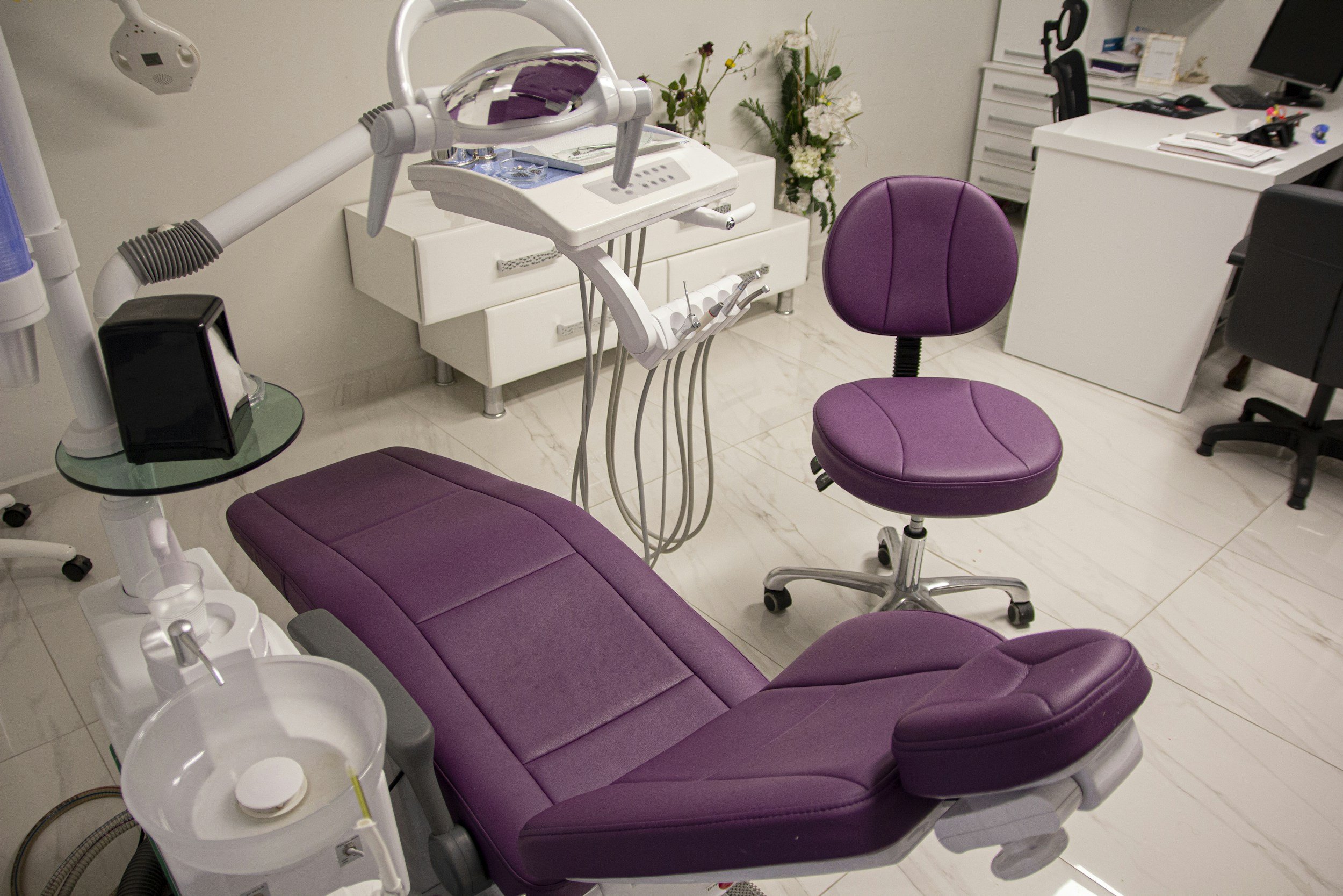 A dental chair in a dental clinic with purple upholstery, surrounded by dental equipment, a white cabinet with flowers, and a desk with a computer and office supplies.