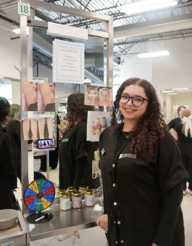 Young woman with glasses in black jacket smiling at a booth with cosmetic or skincare products, colorful informational posters, and a small spinning wheel for a game or giveaway, at an indoor fair or expo.