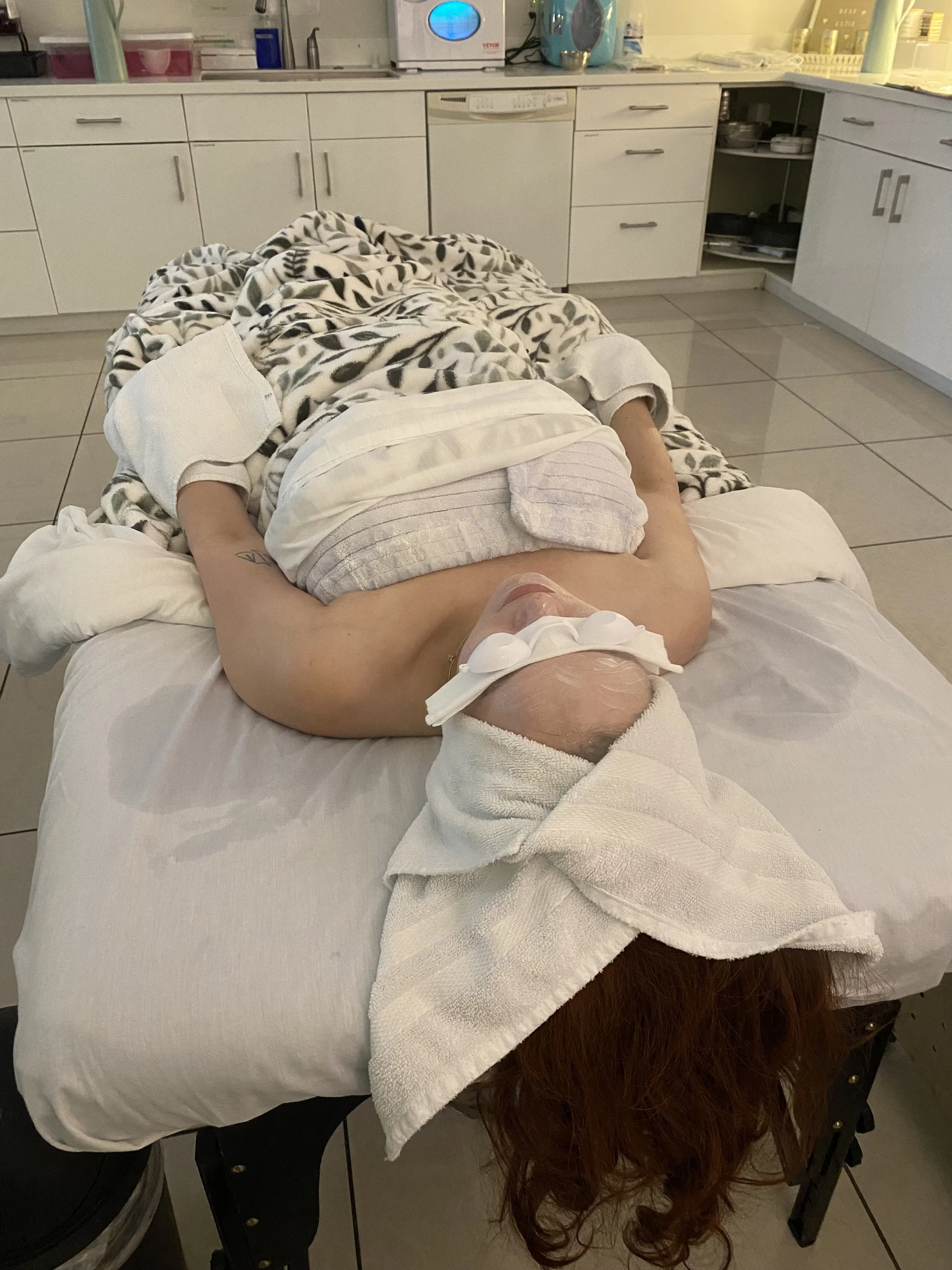 a person lying on a massage table with hair wrapped in a spa towel, eyes covered with goggles, hands wrapped in parrafin wax and covered in towels, steamer on the face 