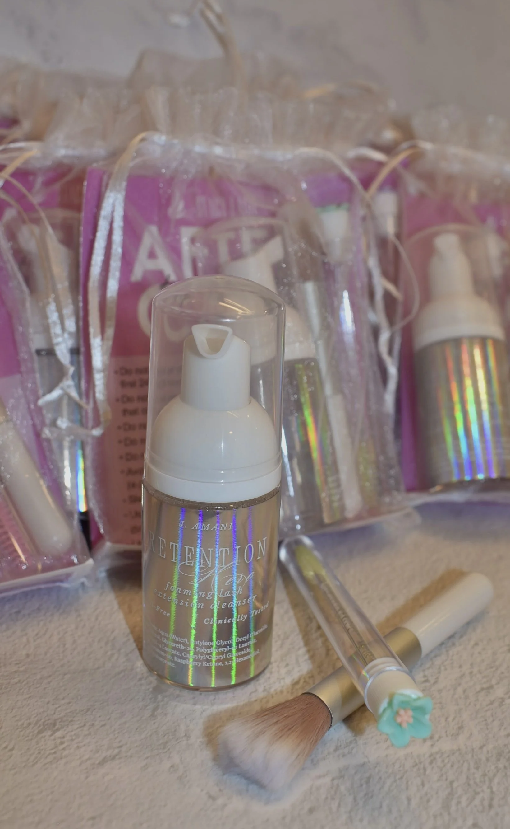 A clear plastic bottle of foaming facial cleanser with a white pump top, a makeup brush with a floral handle, and several small skincare sample packets in pink packaging are arranged on a beige surface.