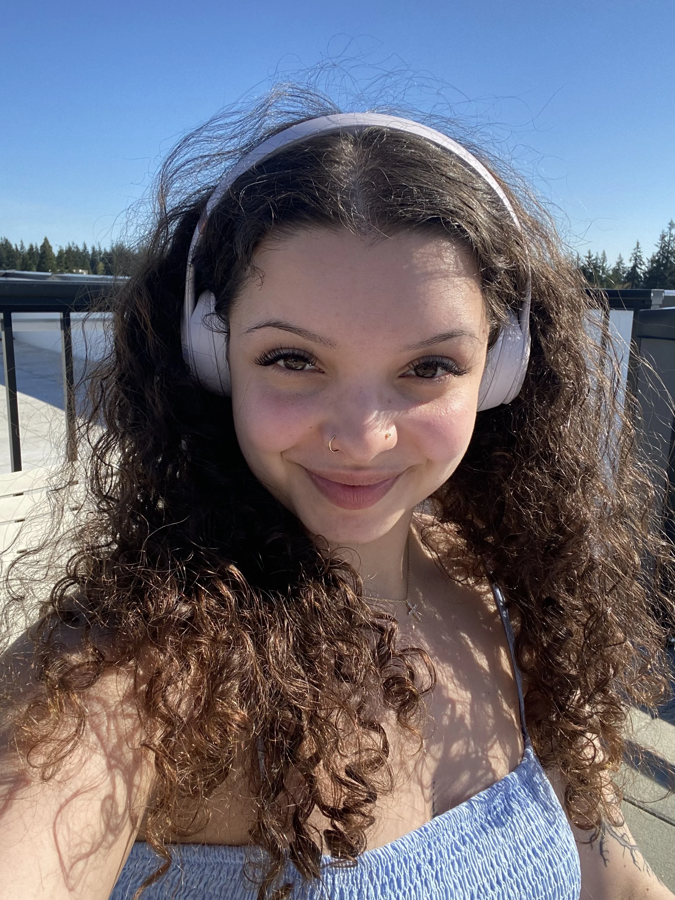 A woman with curly brown hair, wearing white headphones and a blue top, is smiling outdoors against a bright blue sky and some trees.