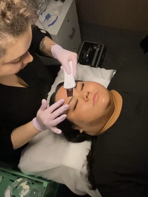 A woman receiving a facial treatment from a skincare professional in a clinical setting, lying on a bed with a pillow, with the professional applying a mask or chemical peel to her face using a tool. The professional is wearing gloves and a black outfit.