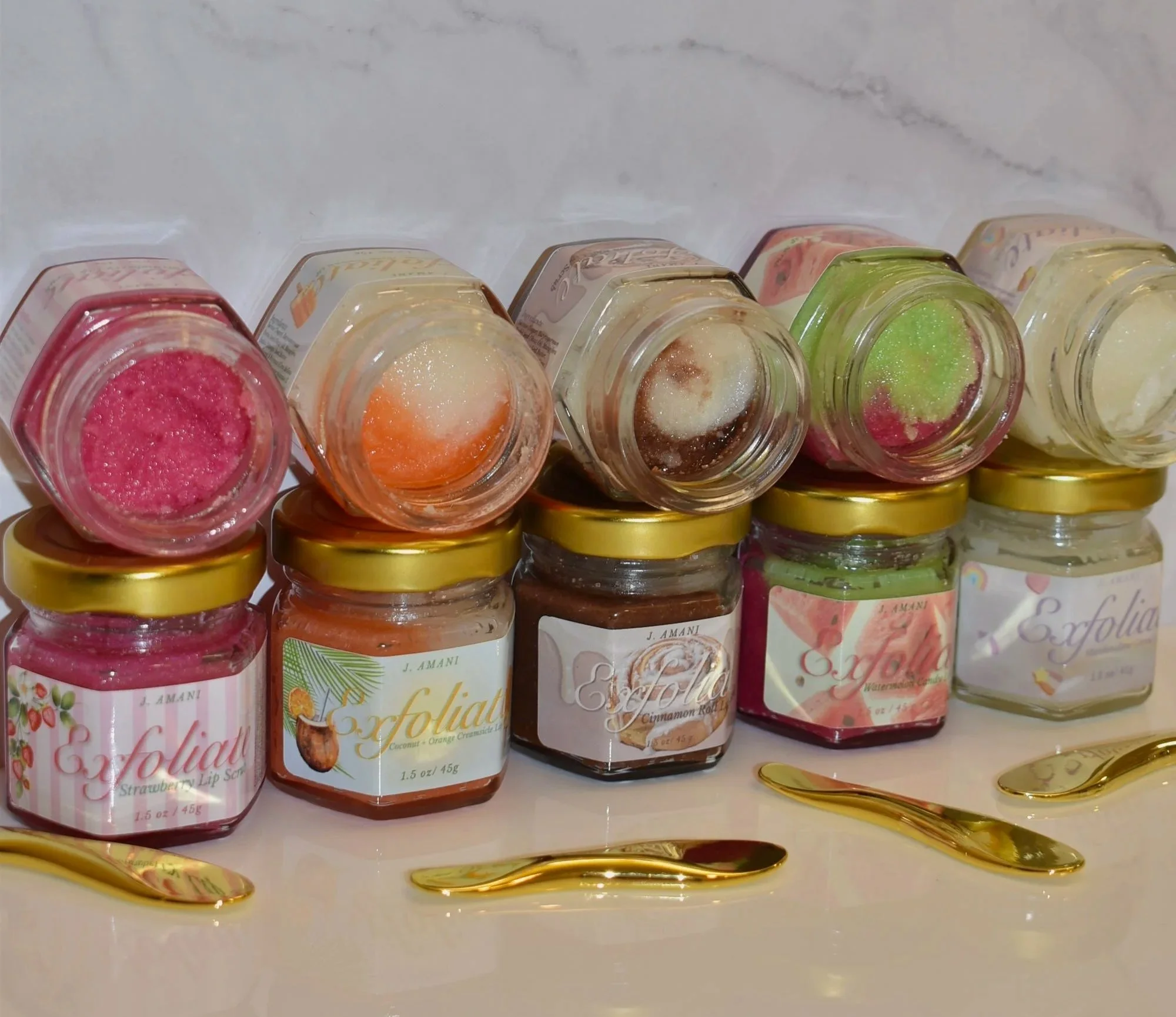 A row of small jars of flavored lip scrubs in various colors, including pink, orange, green, and cream, with gold lids, displayed on a shiny gold tray.