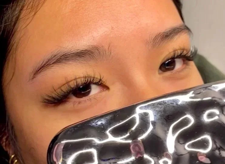 Close-up of a person's face with eyes partially closed, holding a shiny black object reflecting light, obscuring the lower part of the face. Showing an example of hybrid cat eye lash extensions