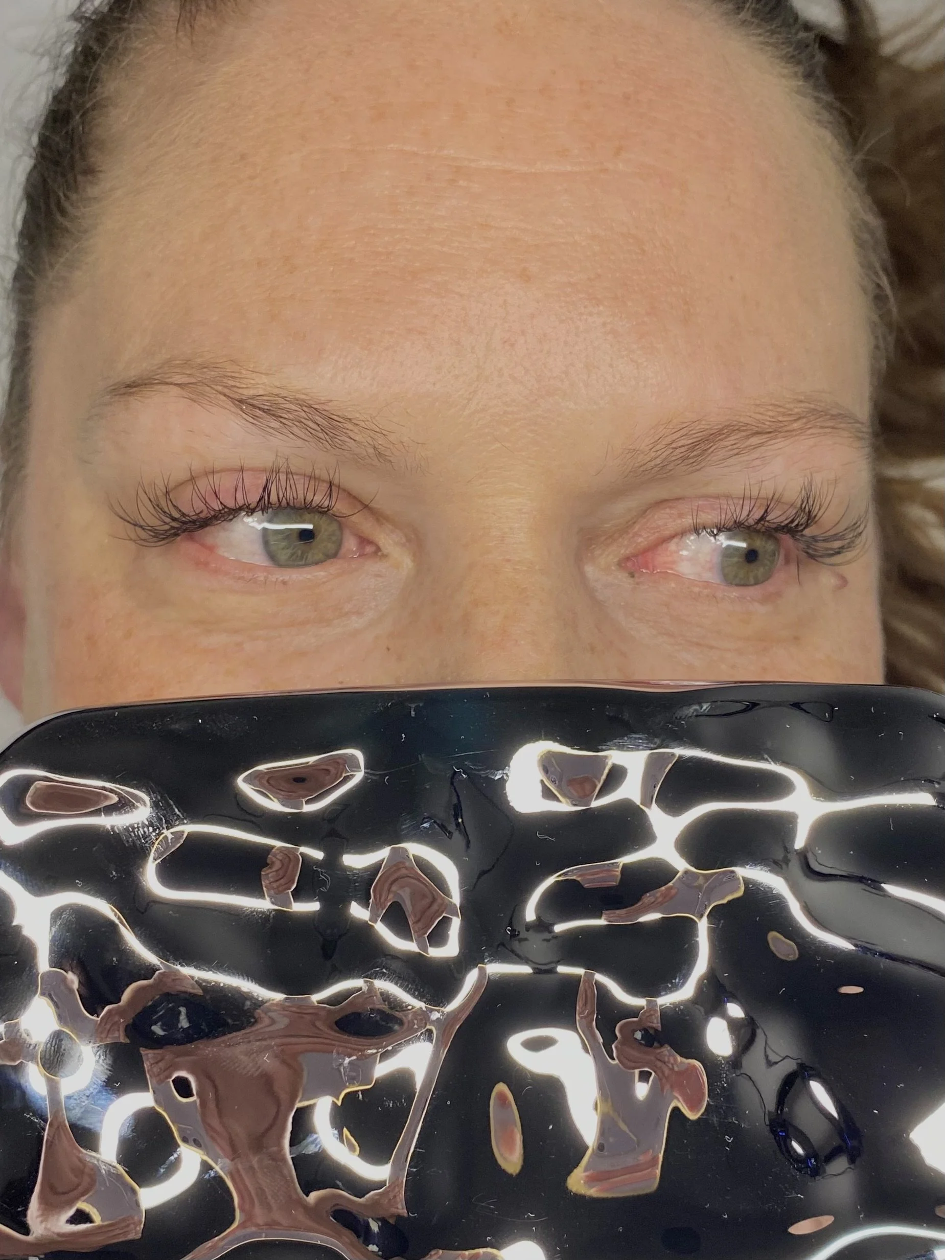 Close-up of a person's face with blue eyes and long eyelashes, showing the face reflected in a shiny, black, abstract surface with irregular, glowing edges. showing classic lash extensions 