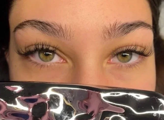 Close-up of a woman's eyes with natural eyebrows, partially covered by a reflective metallic surface. Showing classic lash extensions