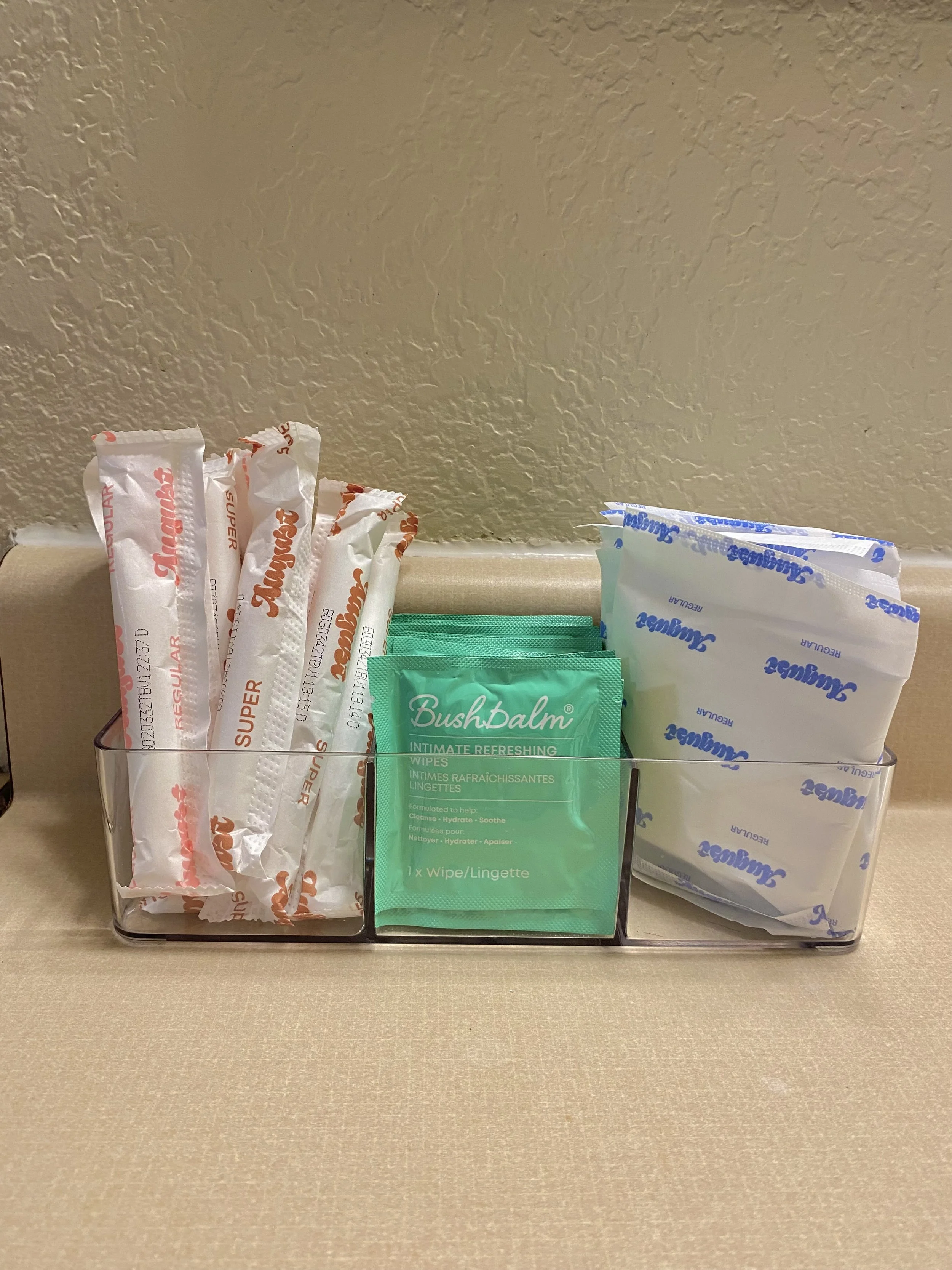 A plastic container  filled with pads, including orange and purple packaging for organic products, with feminine hygiene wipes and tampons as well