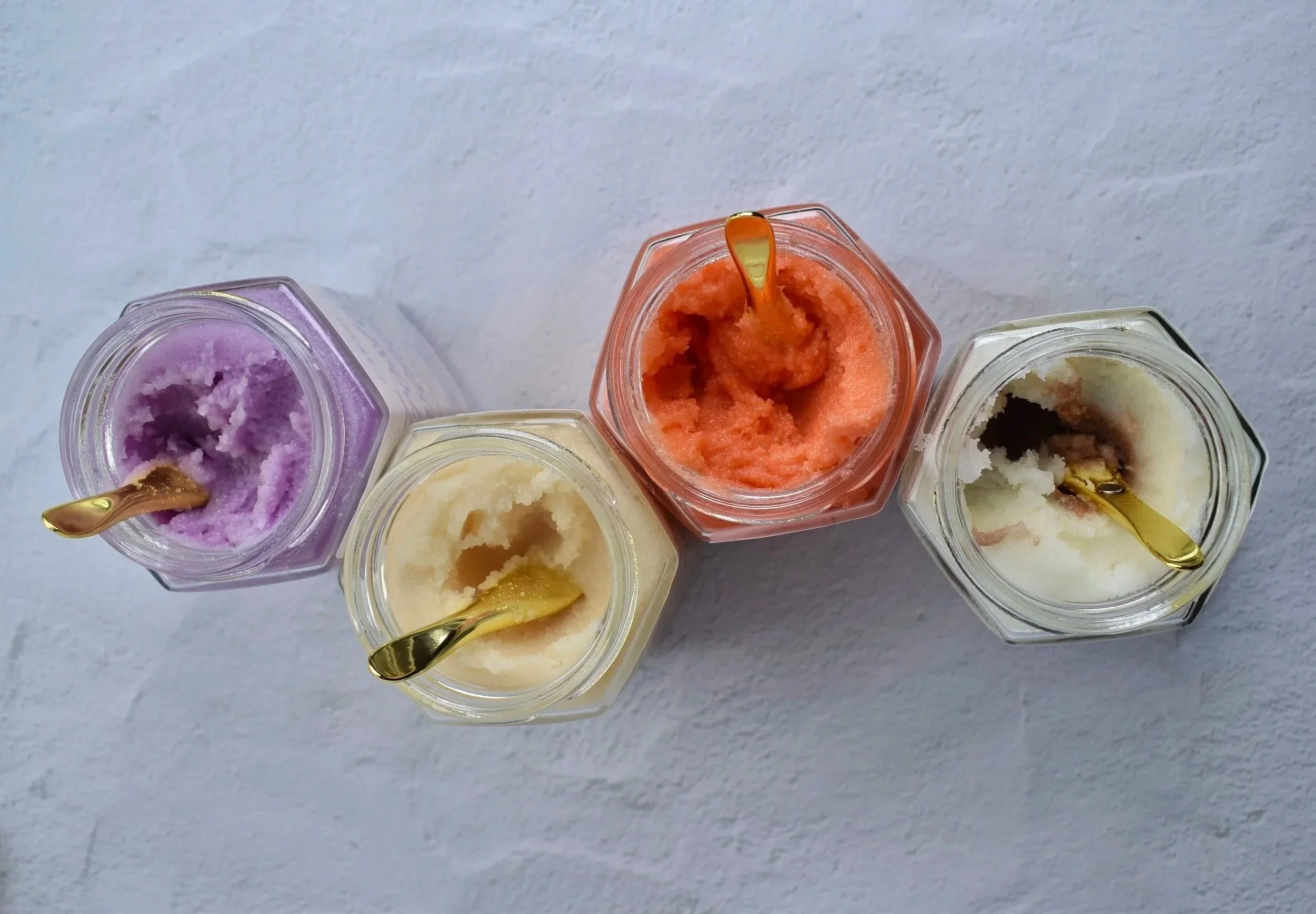 Top view of five small jars of different flavored sorbets with small gold-colored spoons, placed on a white surface.