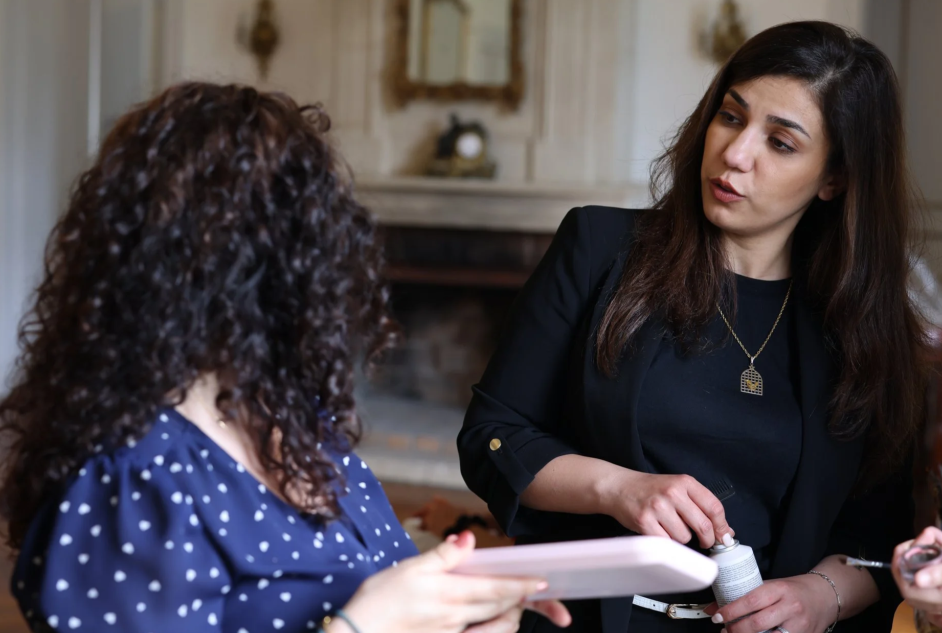 Two women having a conversation indoors, one holding a tablet and the other holding a pill bottle. The woman facing the camera has long dark hair, wearing a black blazer, and a gold necklace. The other woman has curly brown hair and is wearing a blue polka dot blouse.