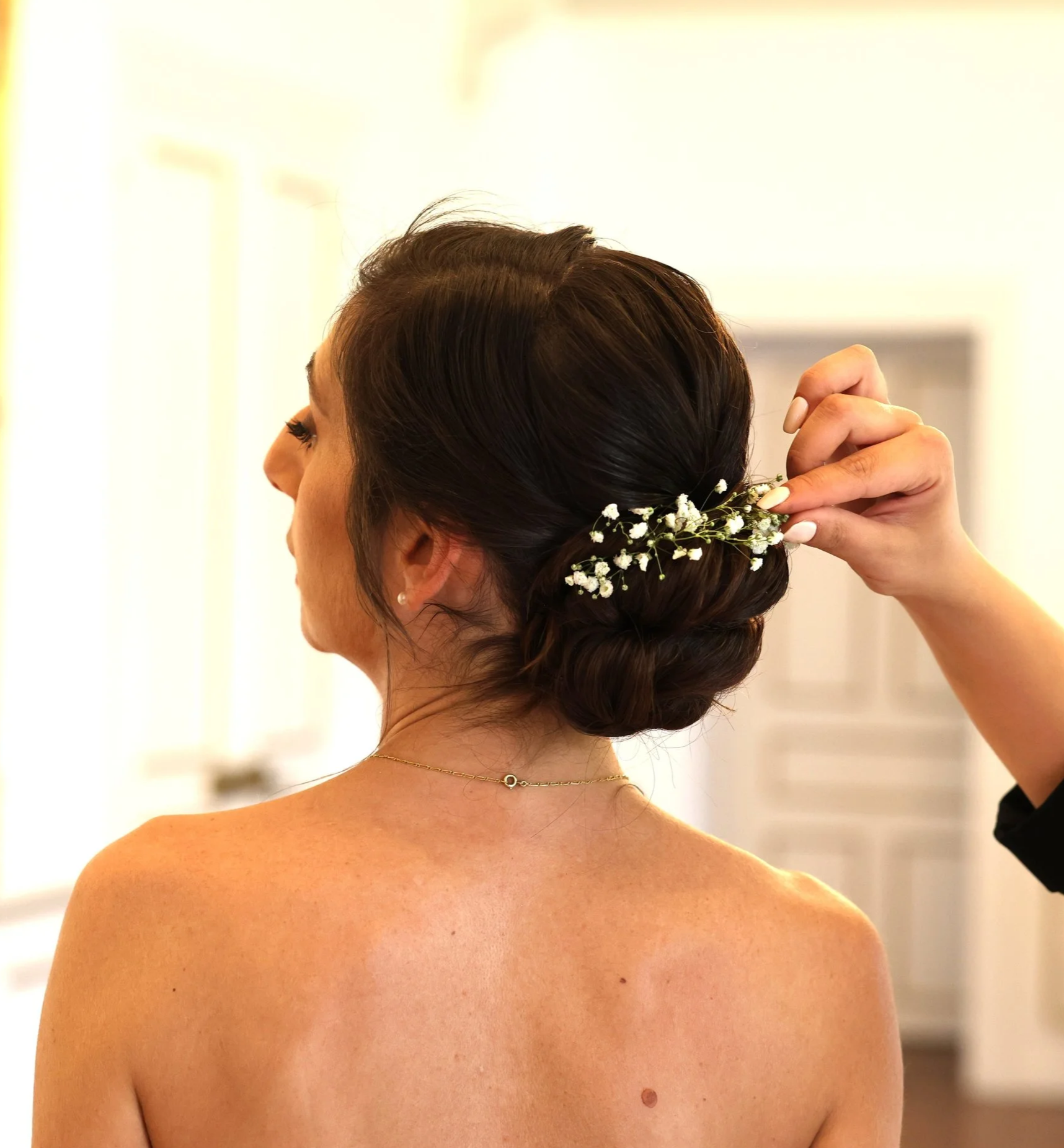 A woman with dark brown hair styled in an elegant updo, adorned with small white flowers, as someone adjusts her hair, and a necklace is visible on her back.