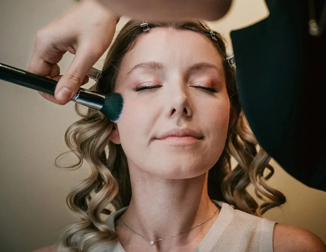 A woman with curled hair and closed eyes having makeup applied with a blush brush by a makeup artist.