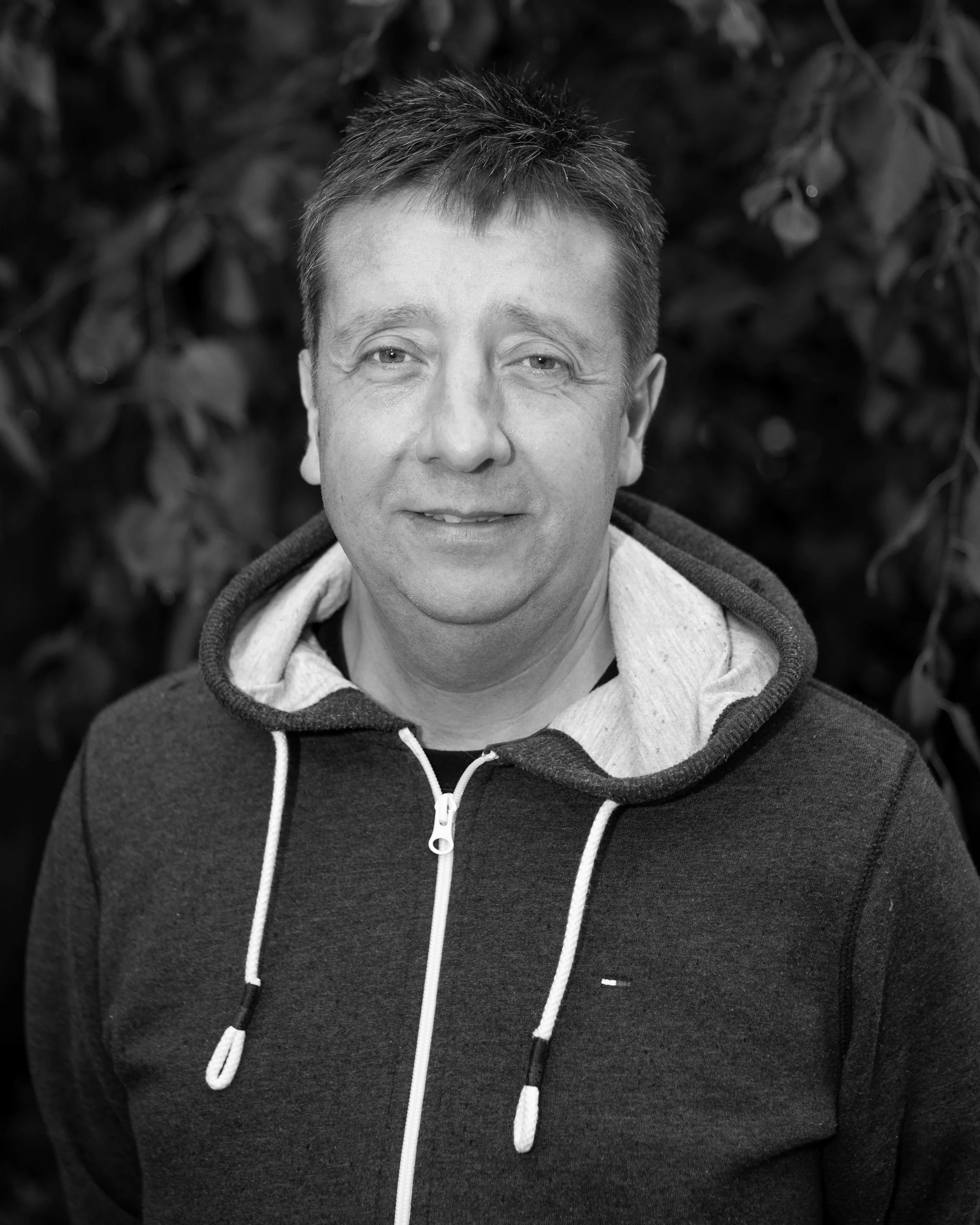 A black and white photo of a smiling man with short hair, wearing a hoodie with a zip, standing outdoors in front of a background of foliage.