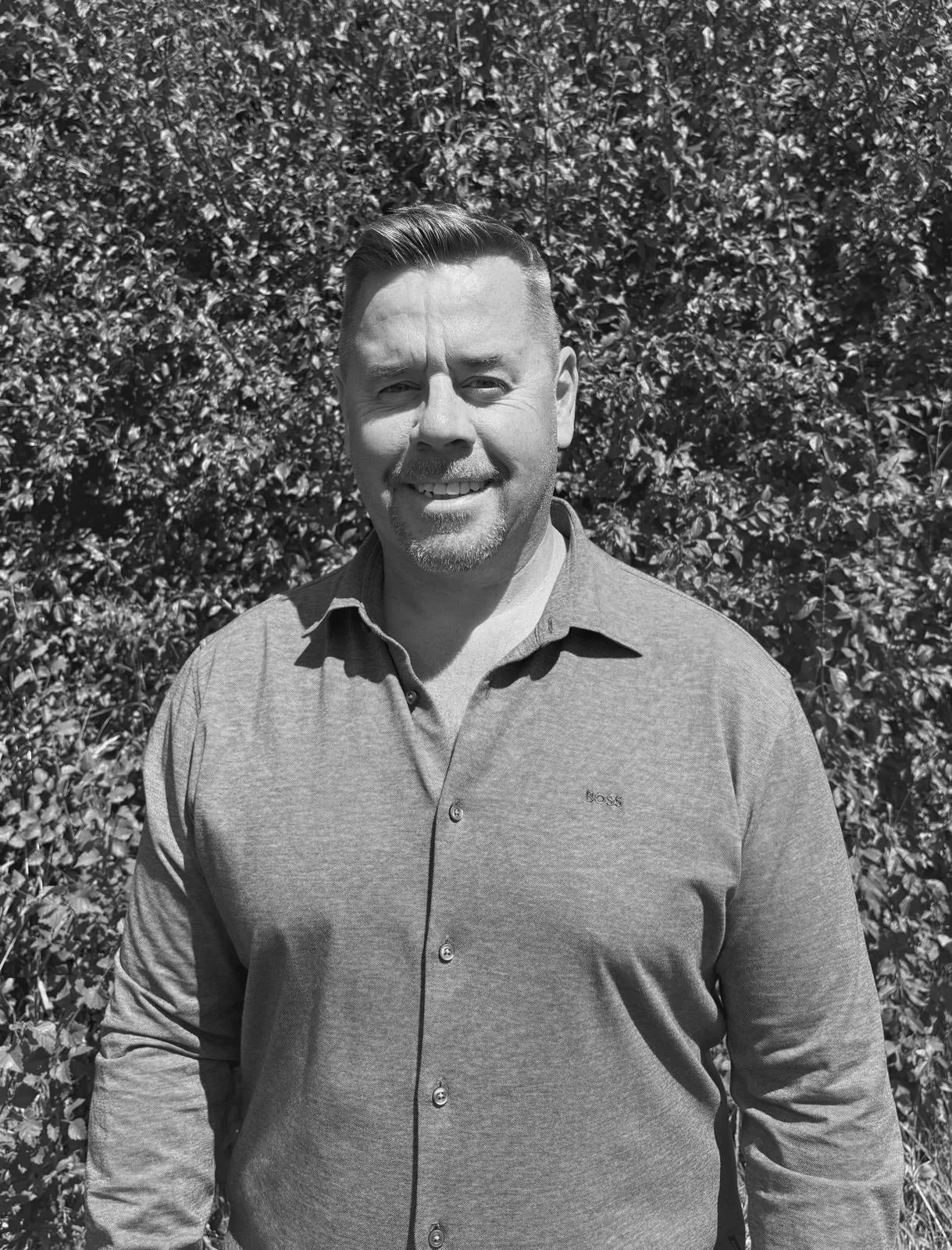 A man smiling outdoors in front of a bush, wearing a button-up shirt.