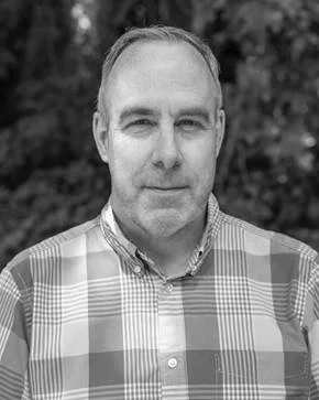 Black and white portrait of a middle-aged man with short hair, wearing a checkered shirt, outdoors with blurred trees in the background.