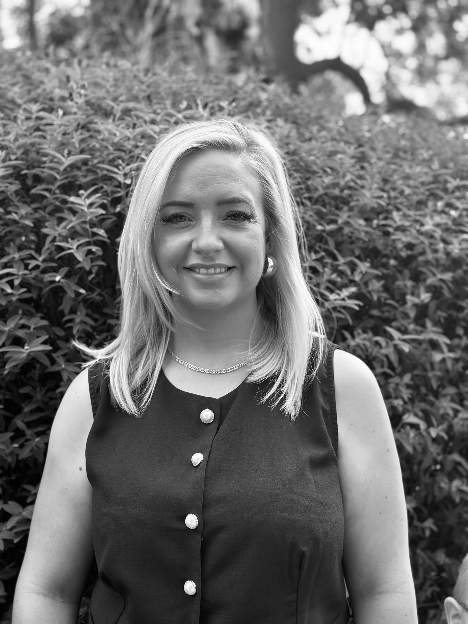 A woman with shoulder-length blonde hair smiling, wearing a sleeveless dark top with pearl buttons, a necklace, and earrings, standing outdoors in front of a bush with trees in the background.