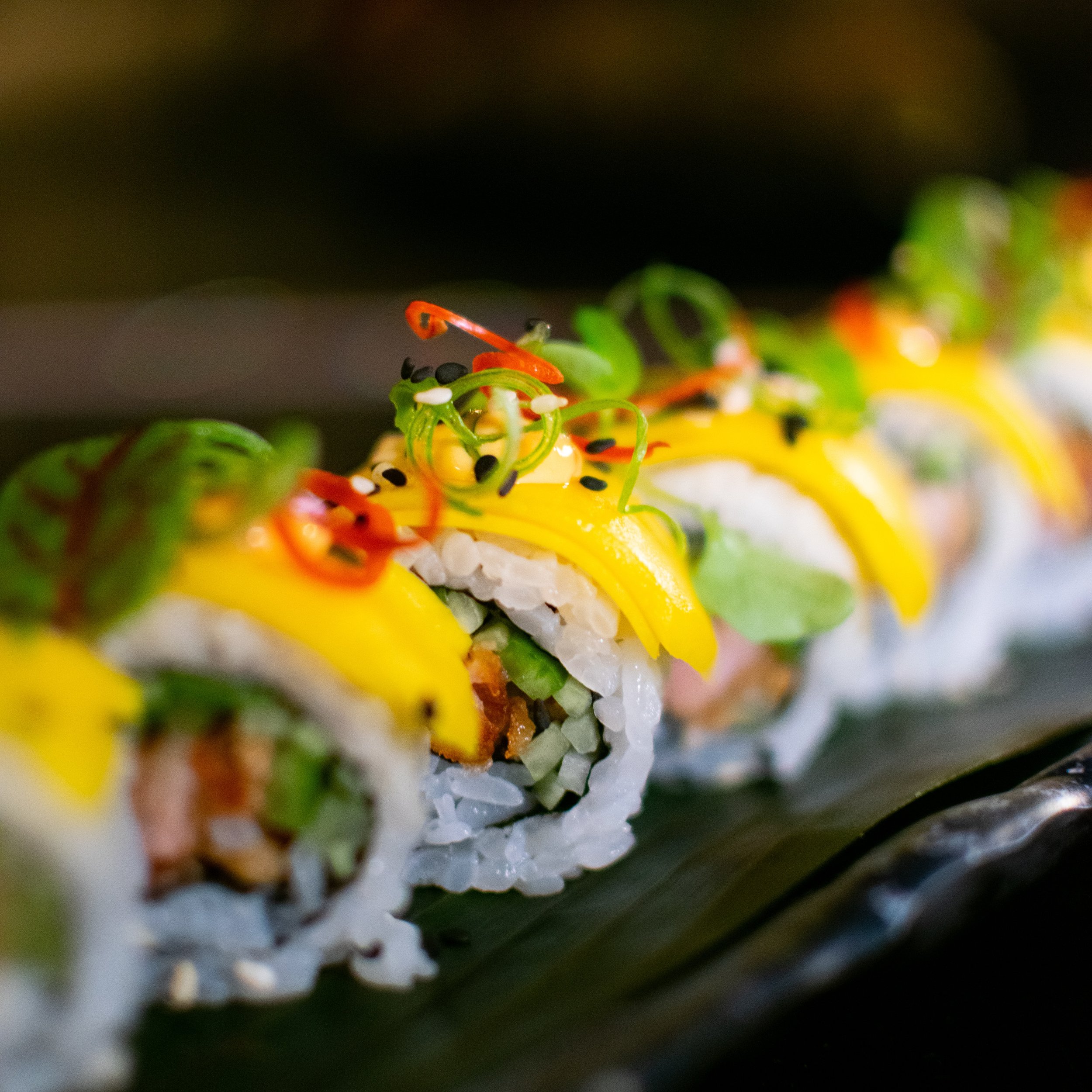 Sushi rolls on a black slate plate with three small dollops of sauce and a small pile of wasabi and ginger. There is a beige teapot in the foreground, a small bowl of rice, and several small plates on a dark table. The background shows a blurred outdoor scene, possibly at a restaurant.