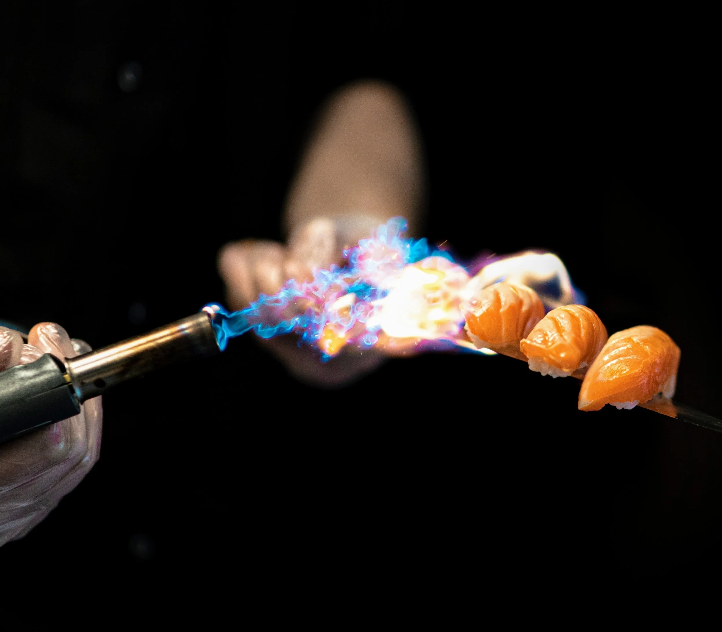A hand holding chopsticks with a piece of sushi against a dark background
