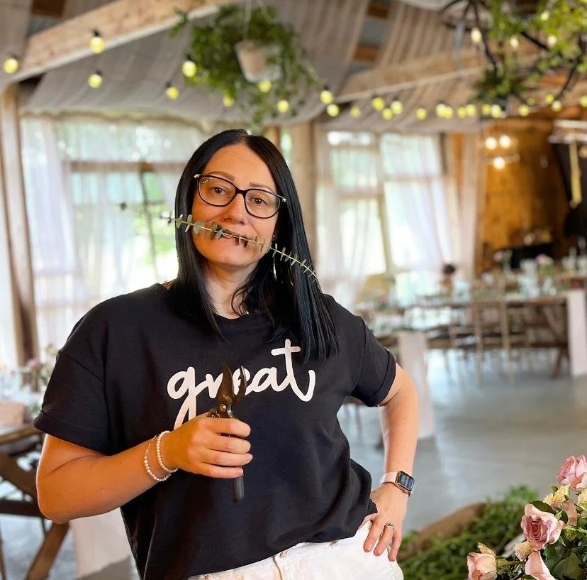 Woman with black hair, glasses, and a black shirt with 'great' written on it, holding gardening tools, standing inside a decorated venue.