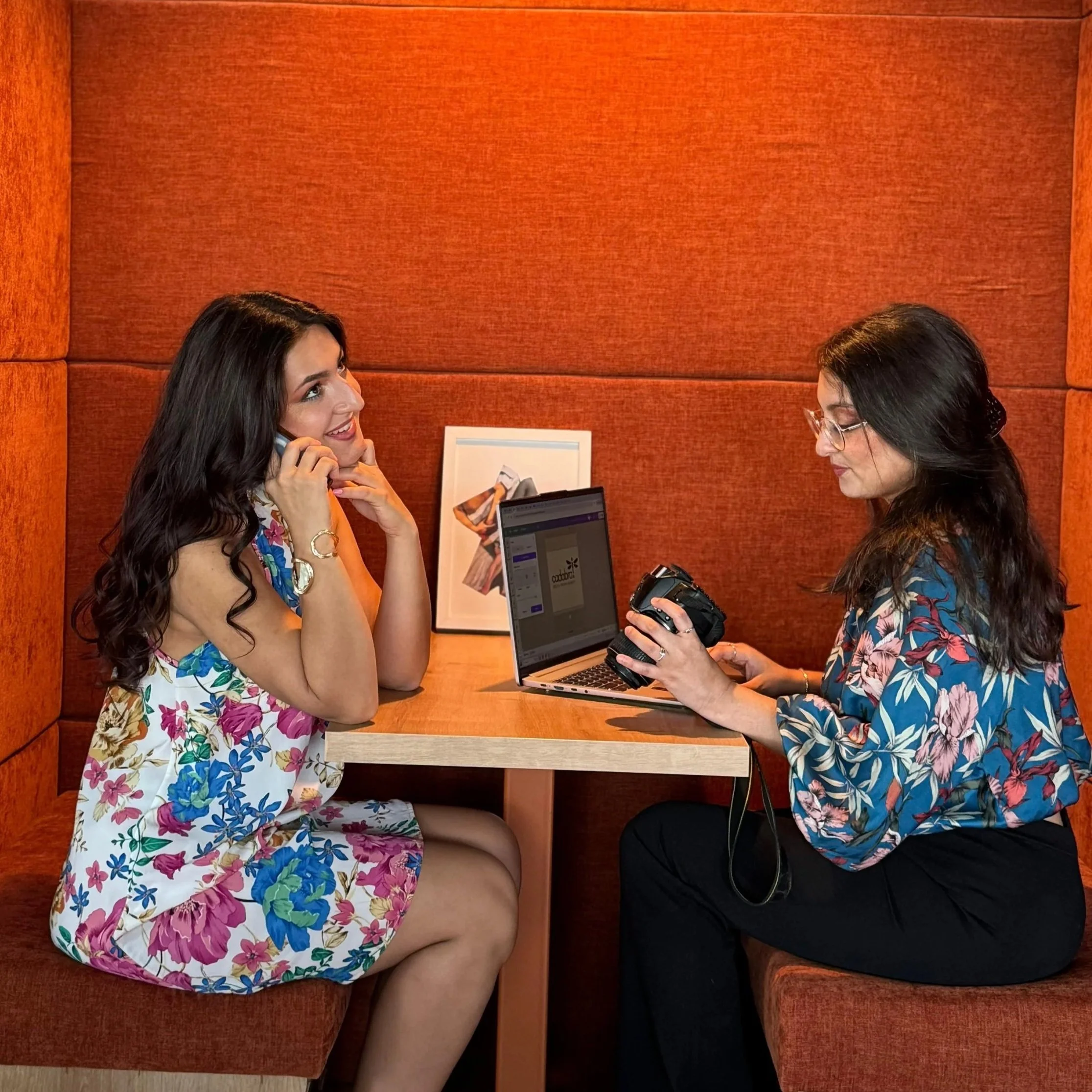Two women sitting at a wooden table in a cozy booth, one talking on the phone and the other holding a camera, with a laptop open between them and a framed picture behind them.