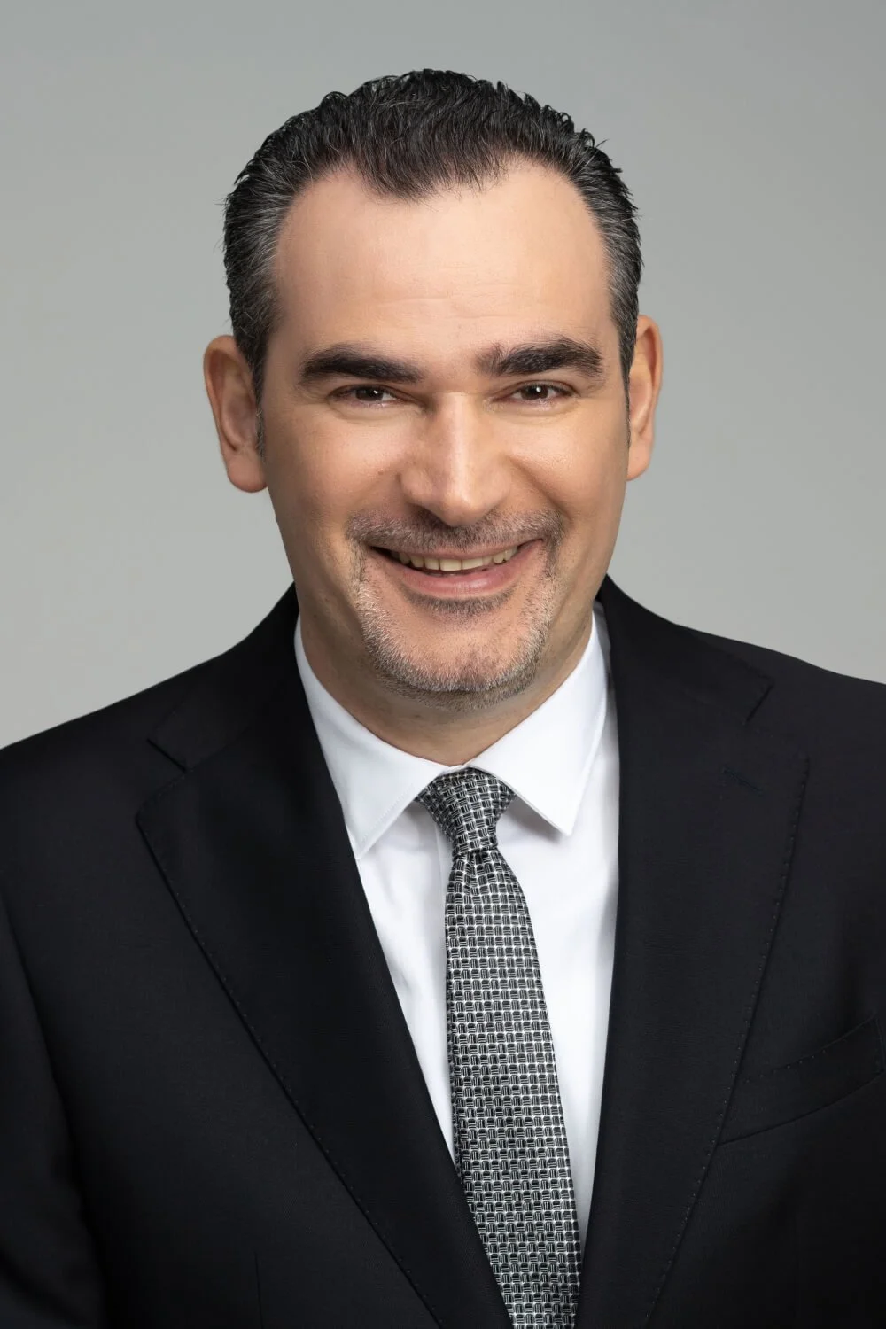 Headshot of a smiling man in a black suit, white shirt, and patterned tie, against a gray background.
