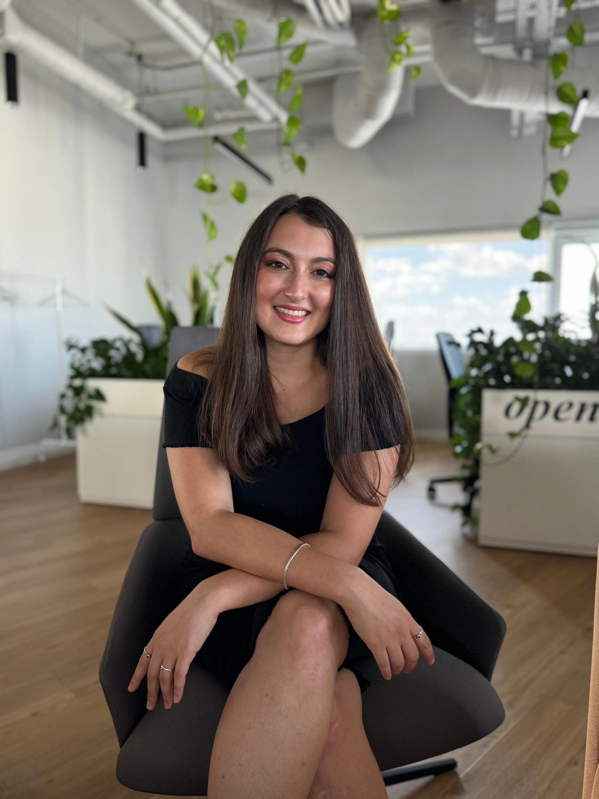 A young woman with long brown hair, wearing a black off-shoulder dress and silver jewelry, sitting on a black office chair in a bright office space with large windows, green plants, and an open sign in the background.