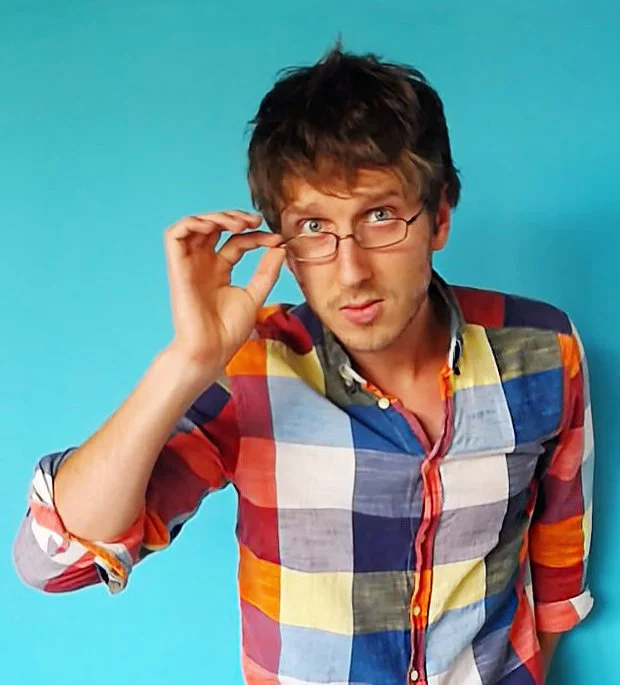 A young man with glasses and tousled brown hair adjusting his glasses while looking at the camera against a bright blue background, wearing a colorful plaid shirt.