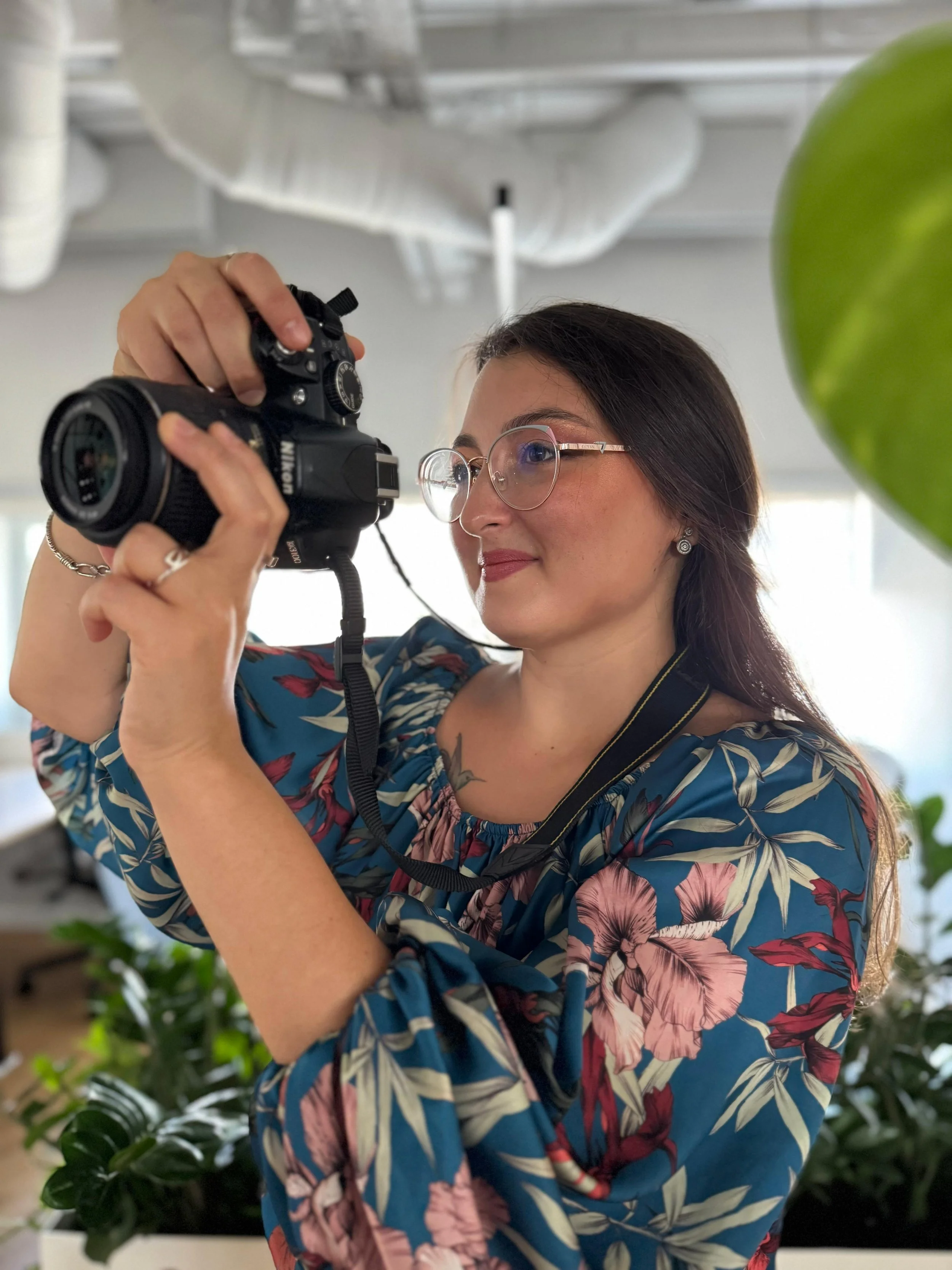 A woman content creator with long dark hair, glasses, and earrings is holding a camera up to her eye in an indoor setting with plants and white ductwork overhead.