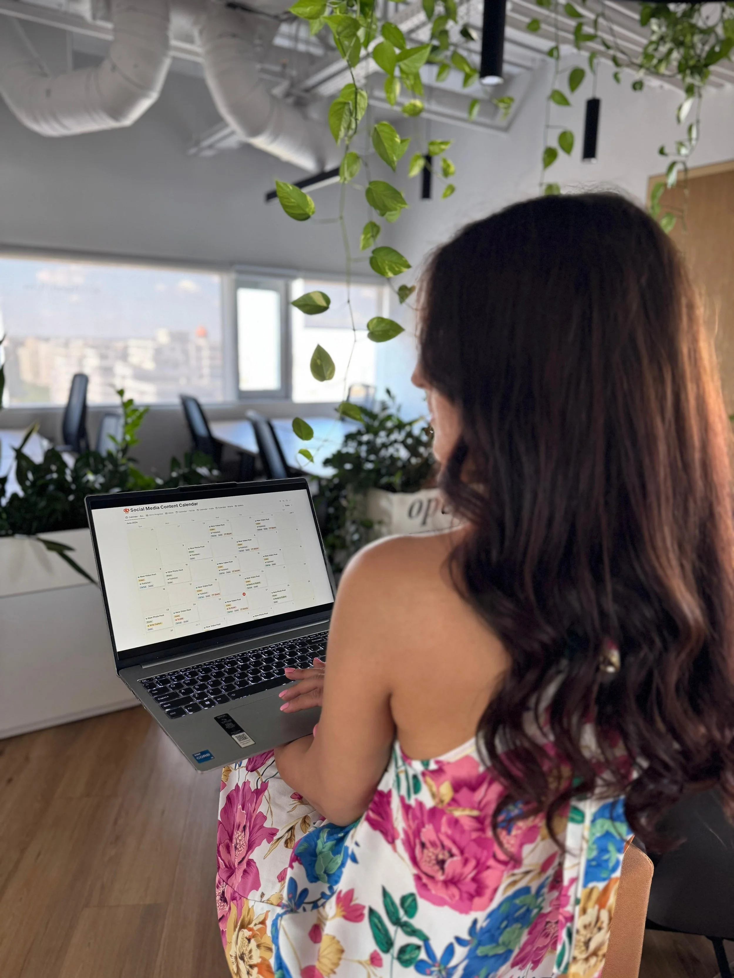 Woman with long hair in a floral dress working on a laptop in a bright office with green plants and large windows on a social media monthly plan
